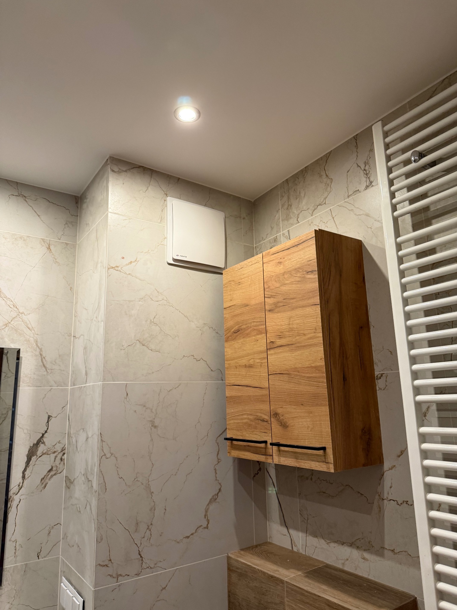 Wykończona łazienka z marmurowymi płytkami, drewnianą szafką pod umywalką i grzejnikiem drabinkowym. Widoczny wentylator i oświetlenie sufitowe.