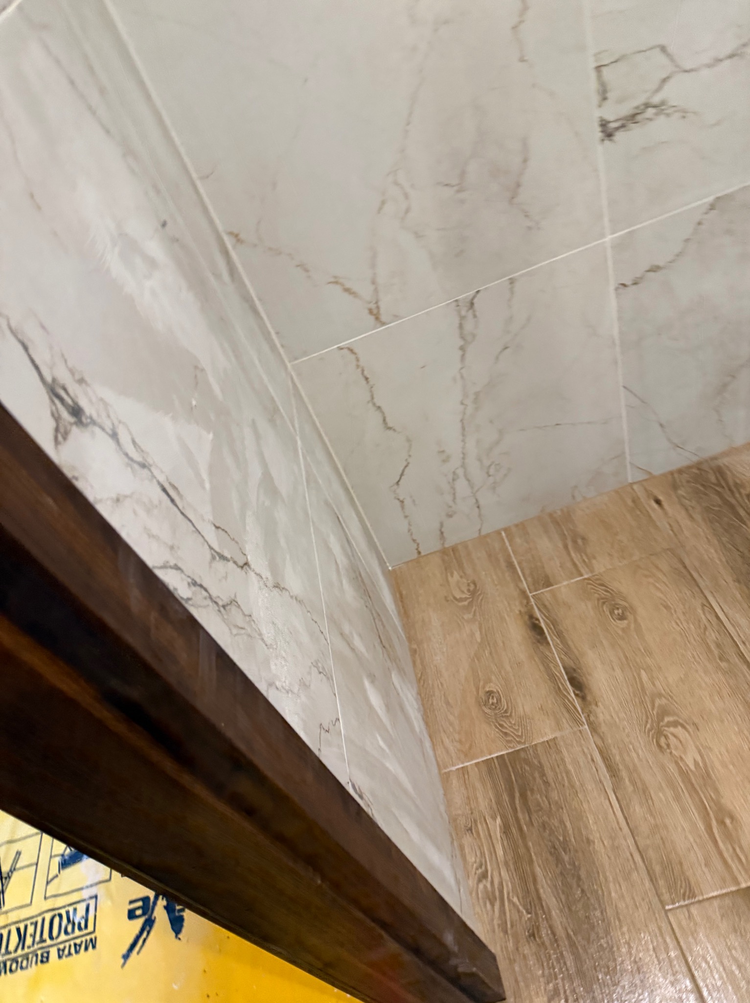 Wykończona łazienka: marmurowe płytki ścienne i podłogowe imitujące drewno. Widoczna krawędź futryny drzwi i fragment folii ochronnej na podłodze.