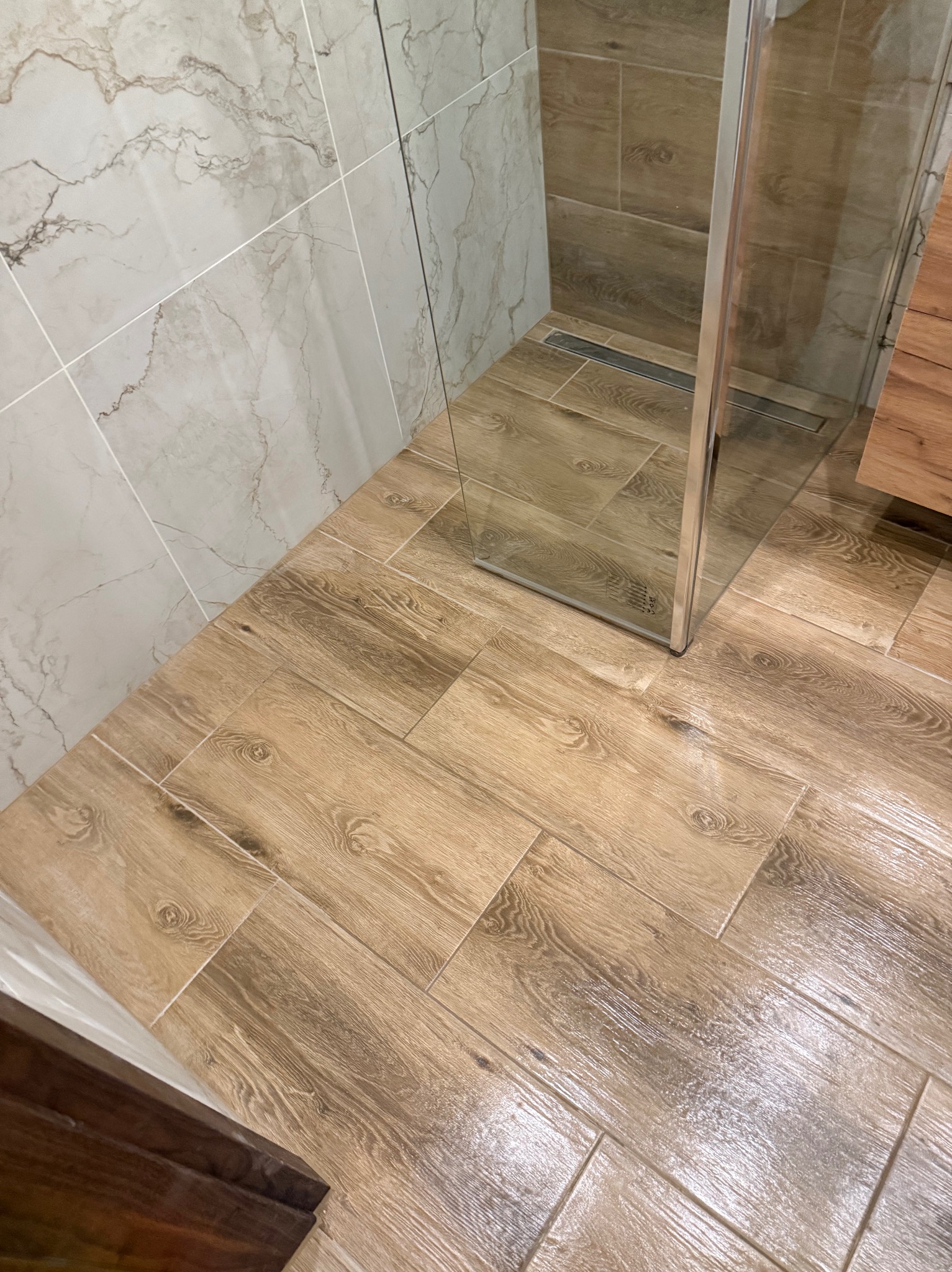 Ukończona łazienka z kabiną prysznicową i drewnopodobnymi płytkami na podłodze ułożonymi w jodełkę. Ściany wyłożone marmurkowymi płytkami.