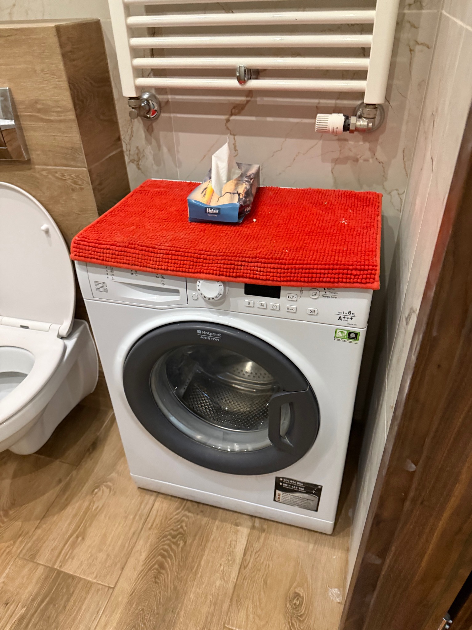 Łazienka z pralką Hotpoint Ariston, na której leży czerwony ręcznik i pudełko chusteczek Velvet. Obok toaleta, na ścianie grzejnik drabinkowy i marmurowe płytki.