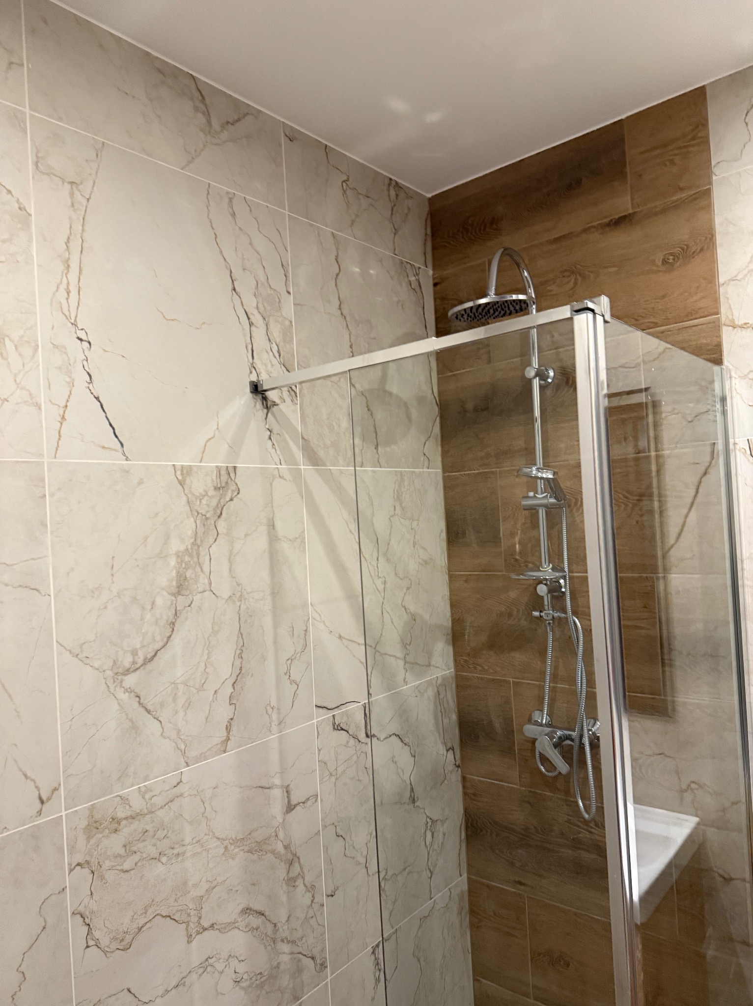 Wykończona kabina prysznicowa z marmurowymi i drewnopodobnymi płytkami w nowoczesnej łazience. Widoczny chromowany zestaw natryskowy i szklane drzwi kabiny.