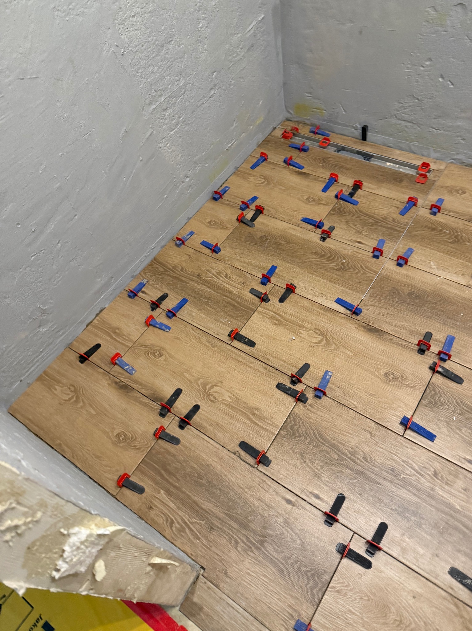 Układanie drewnopodobnych płytek podłogowych w rogu pomieszczenia z użyciem klipsów i klinów poziomujących. Widoczny odpływ liniowy.