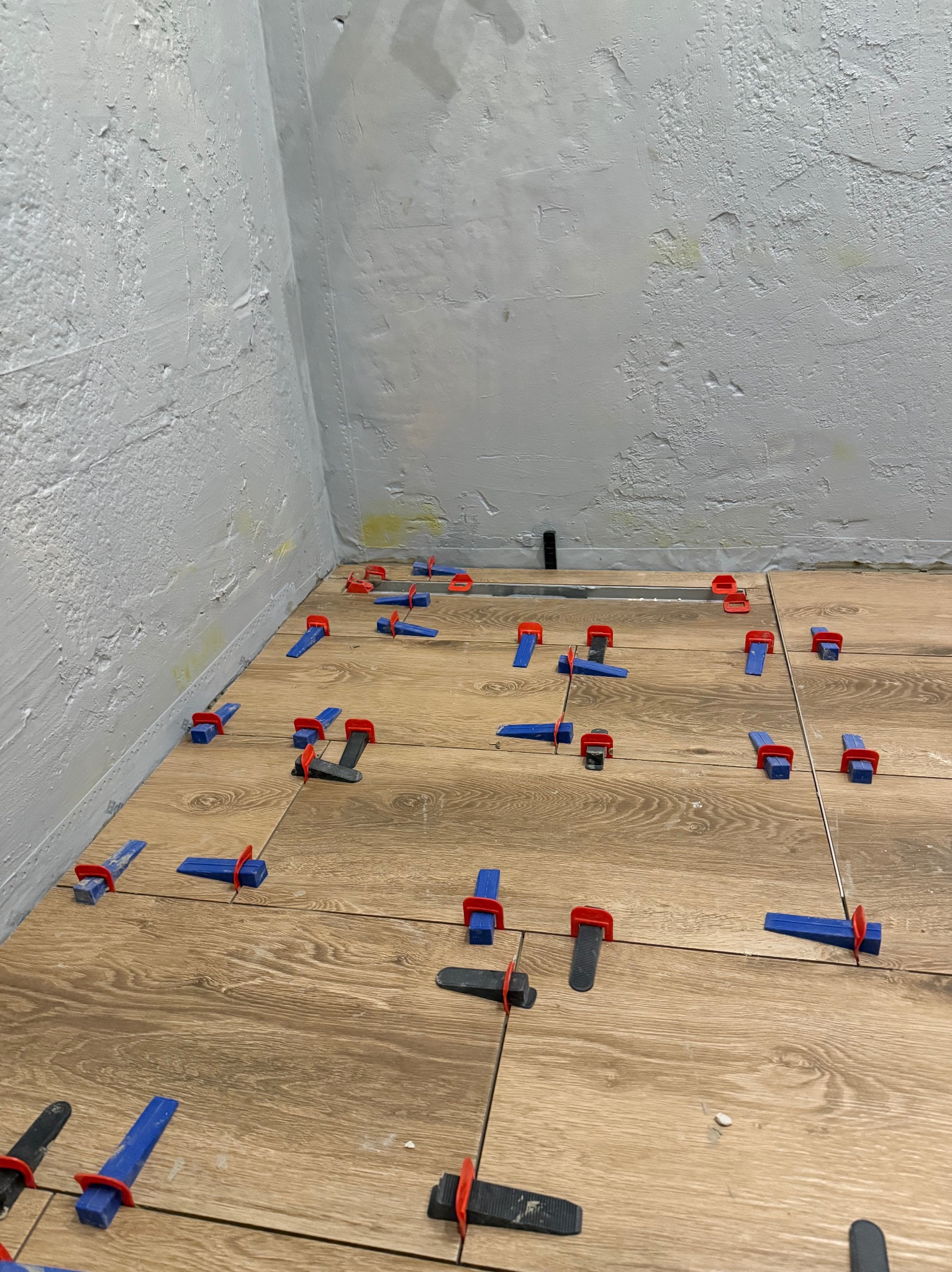 Układanie płytek podłogowych imitujących drewno z użyciem klipsów poziomujących w rogu pomieszczenia z surową, szarą ścianą.