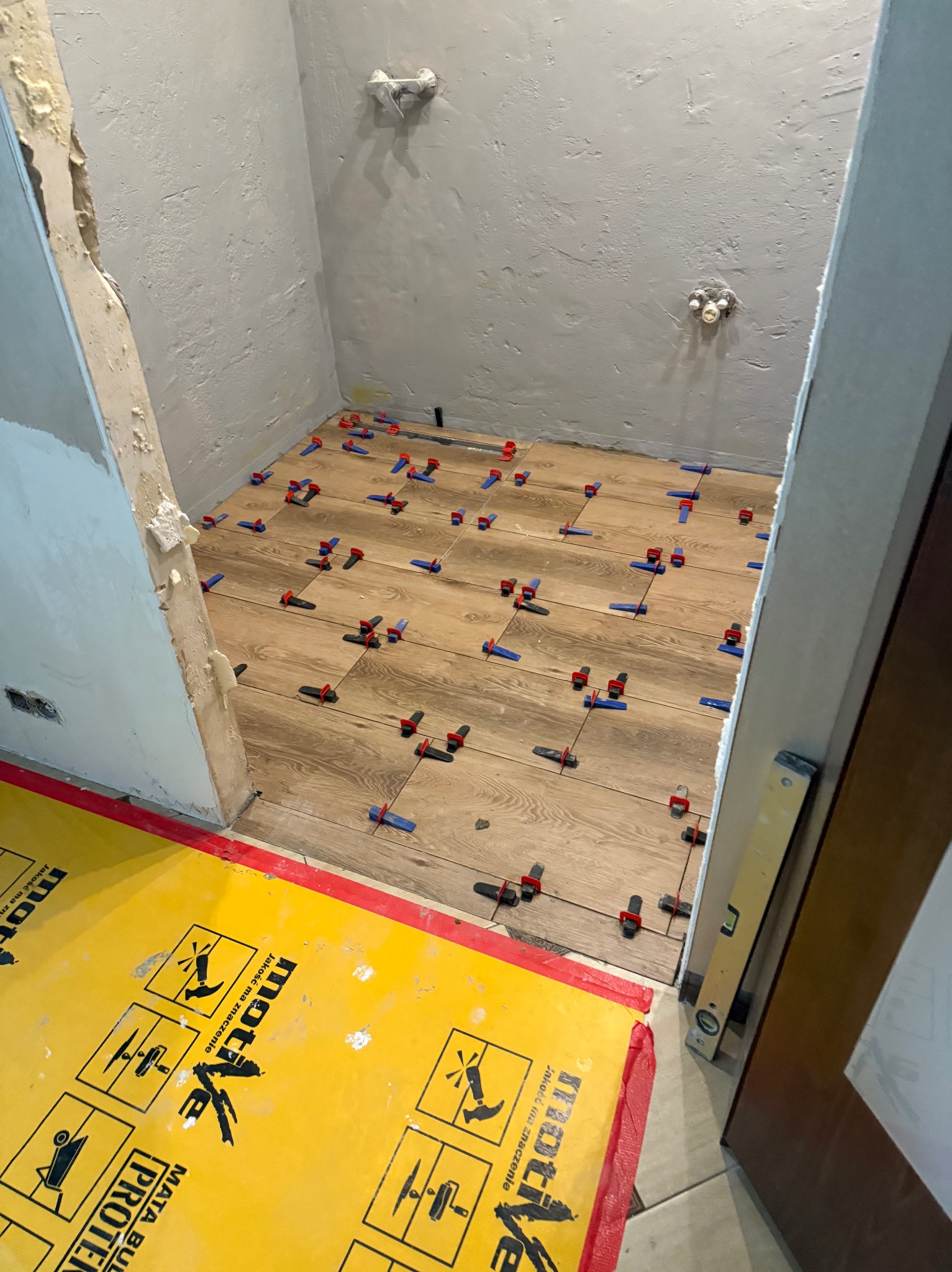 Układanie drewnopodobnych płytek podłogowych w łazience z użyciem systemów poziomowania. Widoczna mata ochronna i poziomica.