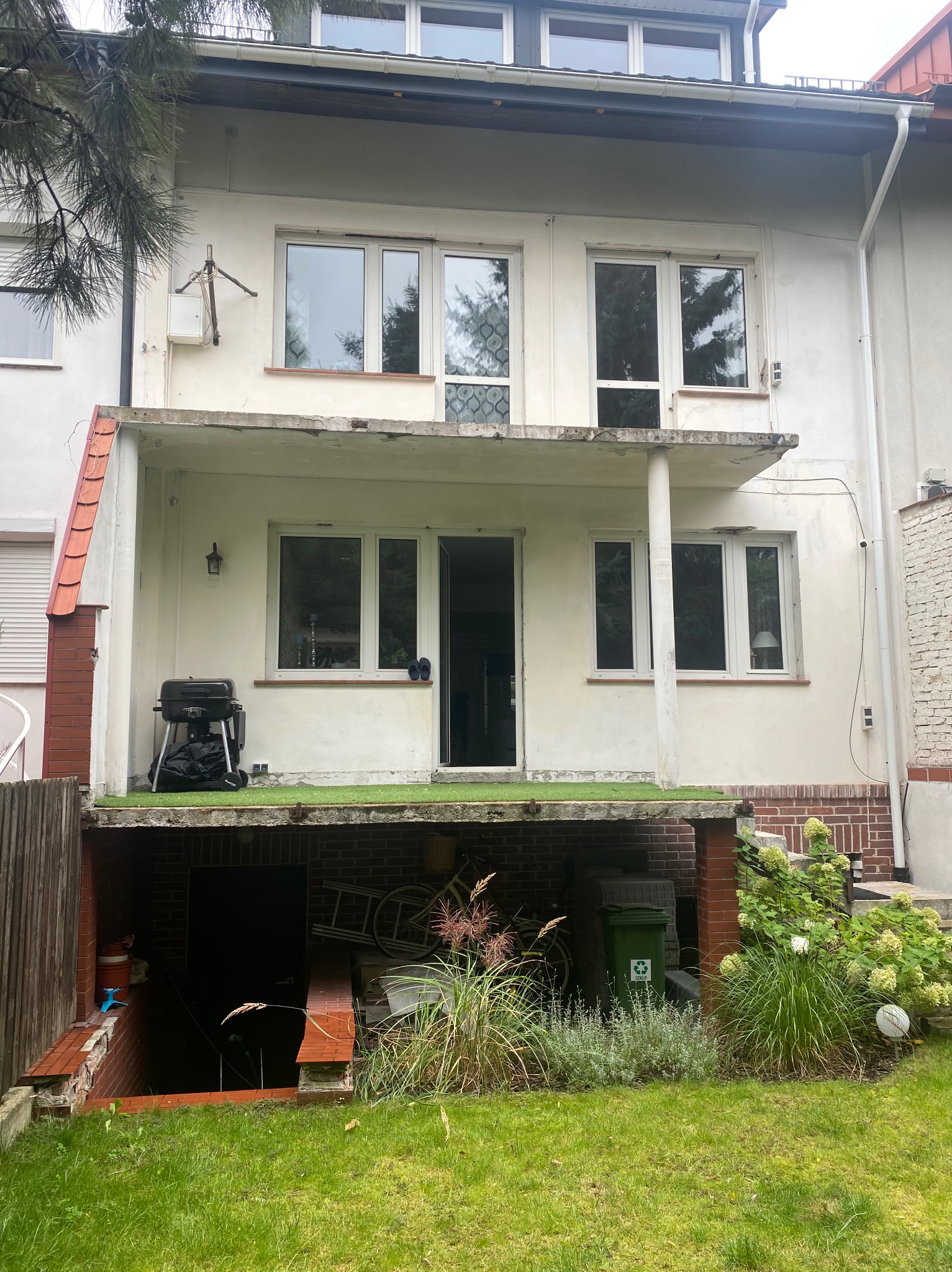 Dwukondygnacyjny budynek z balkonem i zejściem do piwnicy, widok z ogrodu. Na balkonie grill, w piwnicy rower i inne przedmioty. Widoczne okna i elementy wykończeniowe.
