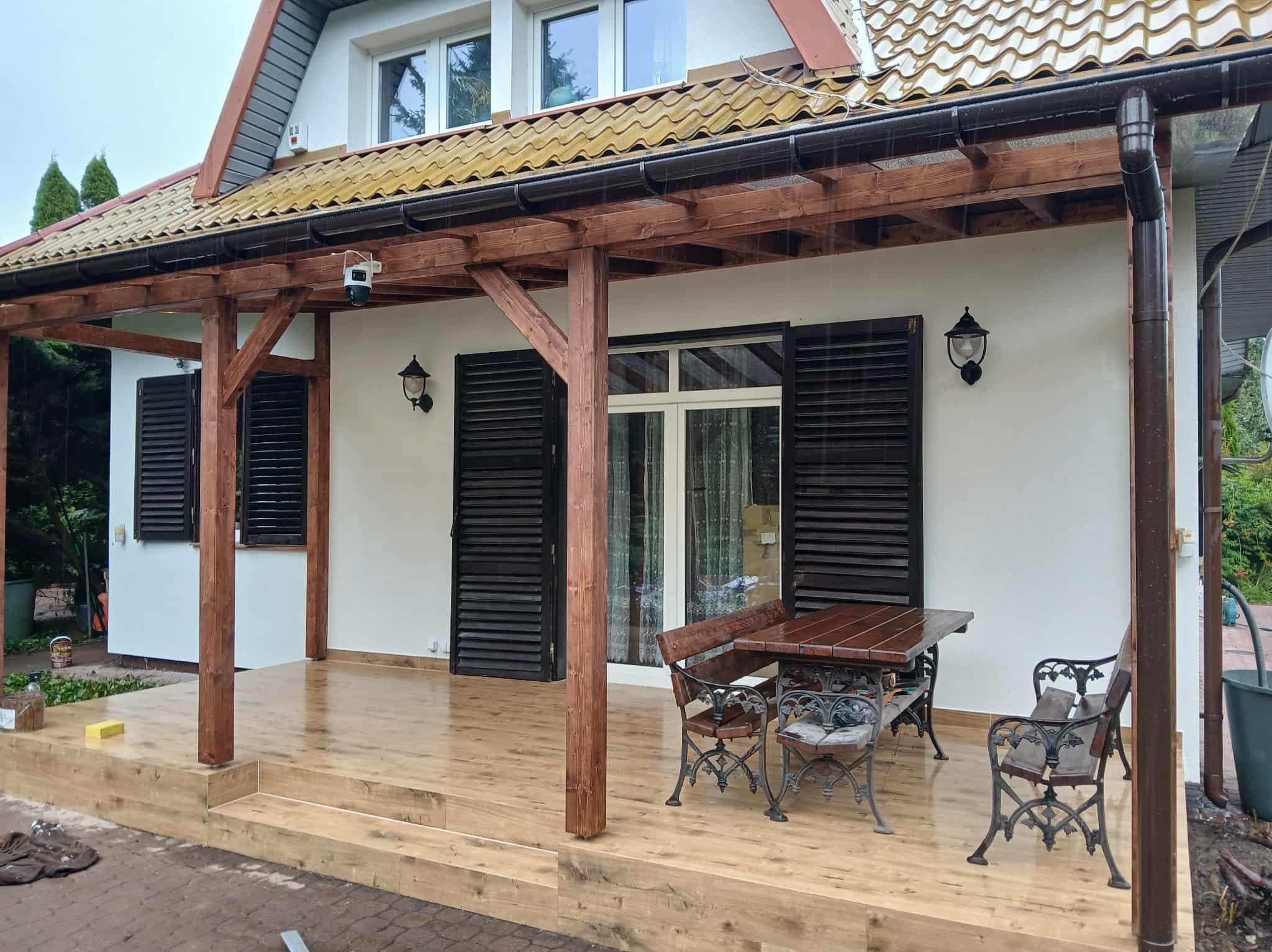 Zadaszony taras z drewnianą podłogą i meblami ogrodowymi. Biała elewacja domu z brązowymi okiennicami i oświetleniem zewnętrznym. Widok na zewnątrz w deszczowy dzień.