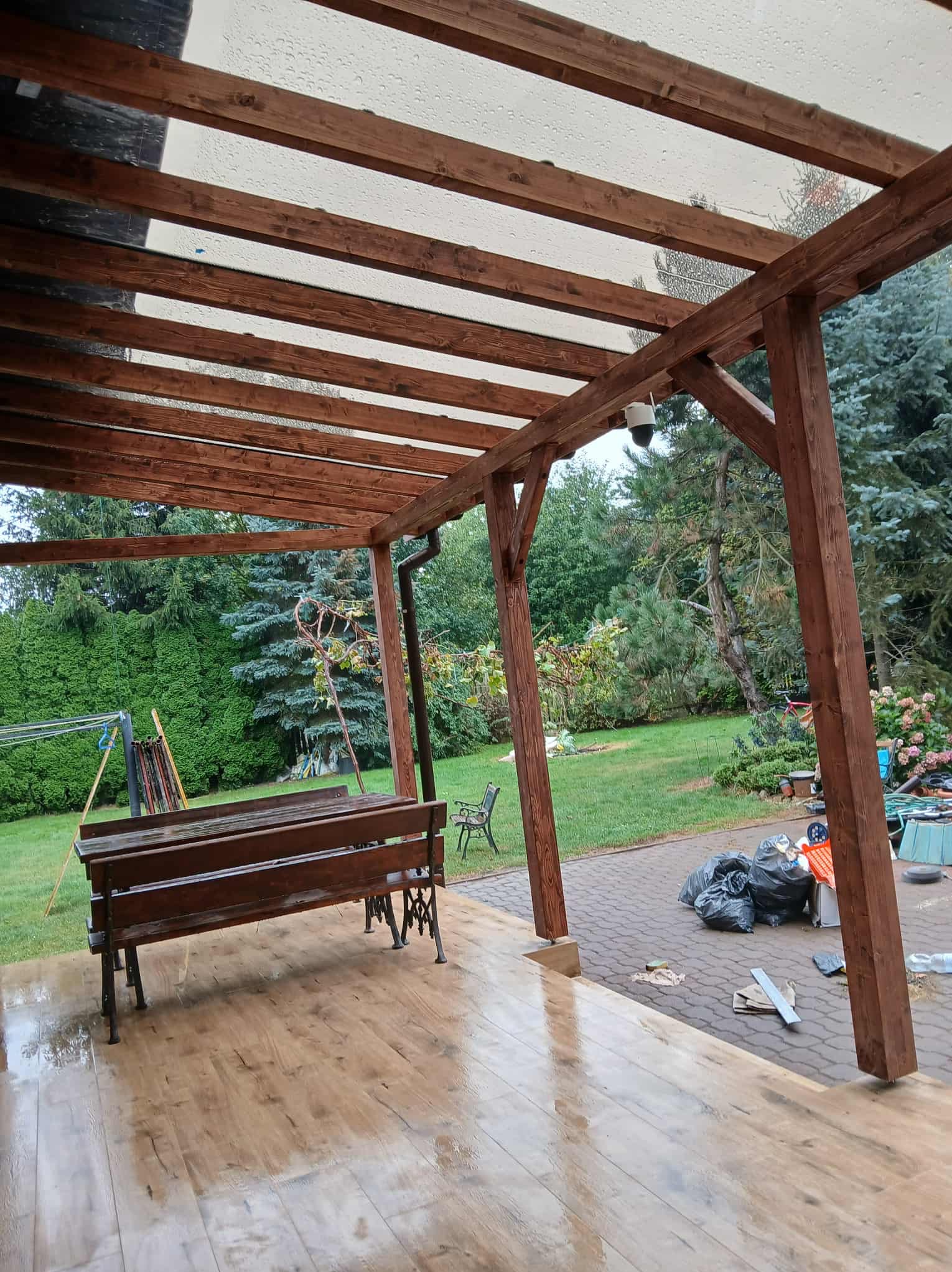 Zadaszona drewniana altana z przezroczystym dachem, drewnianą ławką i widokiem na zielony ogród. Widoczne ślady po deszczu na podłodze i dachu.