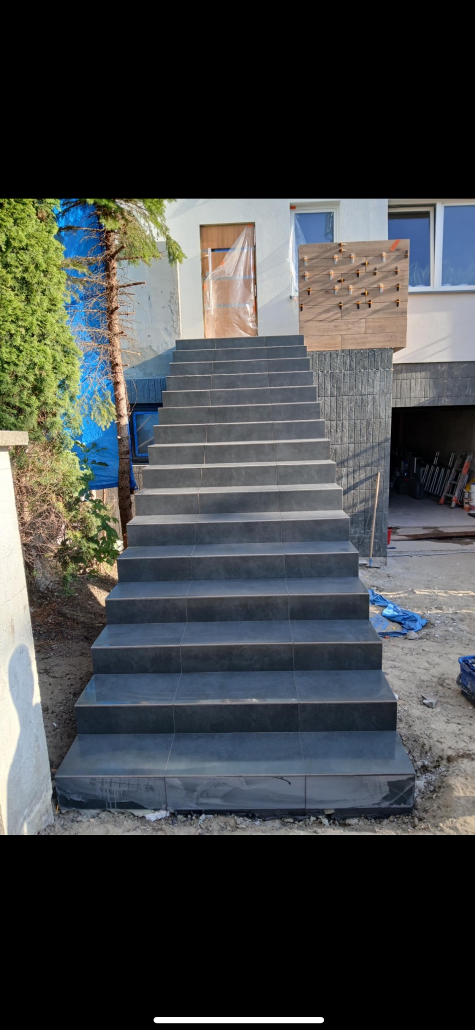 Zewnętrzne schody wyłożone ciemnymi płytkami prowadzące do domu. Widoczne zabezpieczenia na drzwiach i elewacji, co sugeruje trwający proces wykończeniowy.