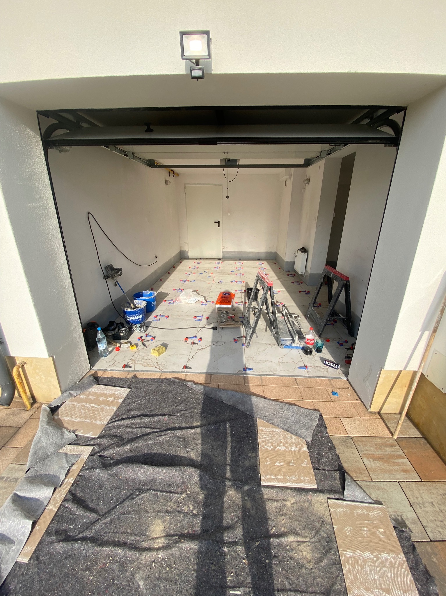 Wnętrze garażu w trakcie układania płytek podłogowych. Widoczne narzędzia, drabina i materiały budowlane. Część płytek już ułożona, część jeszcze nie.