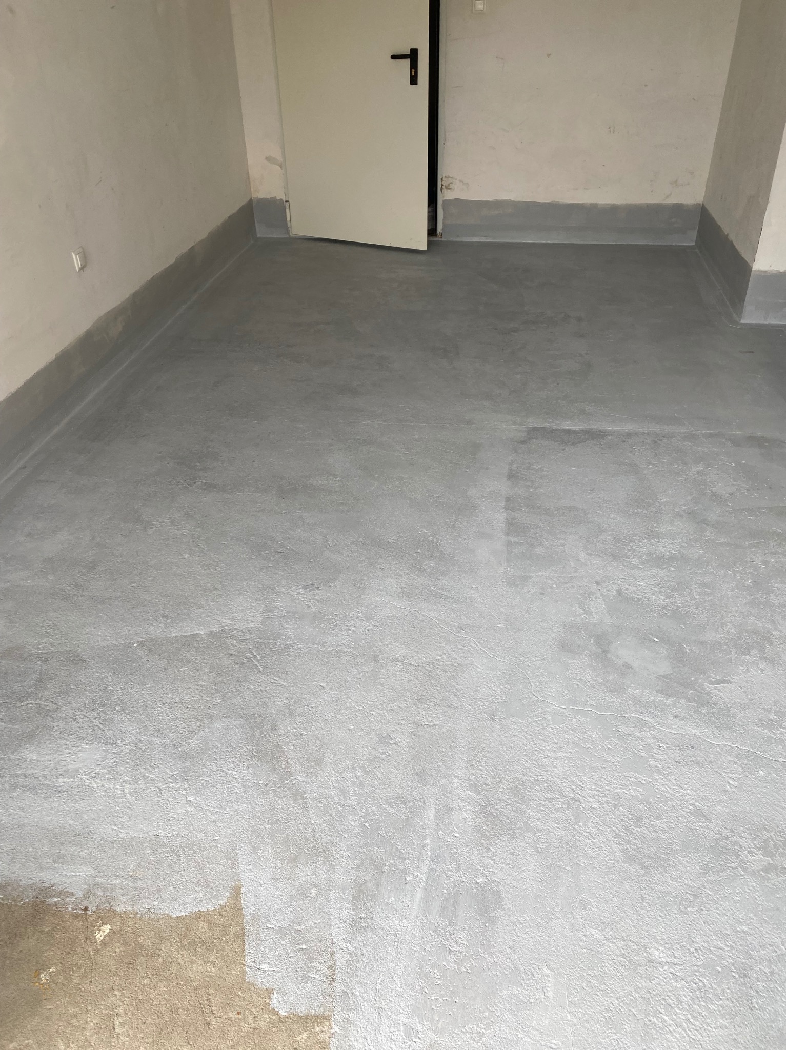 Szara, świeżo wylana posadzka w pomieszczeniu z pomalowanymi ścianami i listwami przypodłogowymi. Fragment surowej podłogi w rogu. Białe drzwi z czarną klamką.