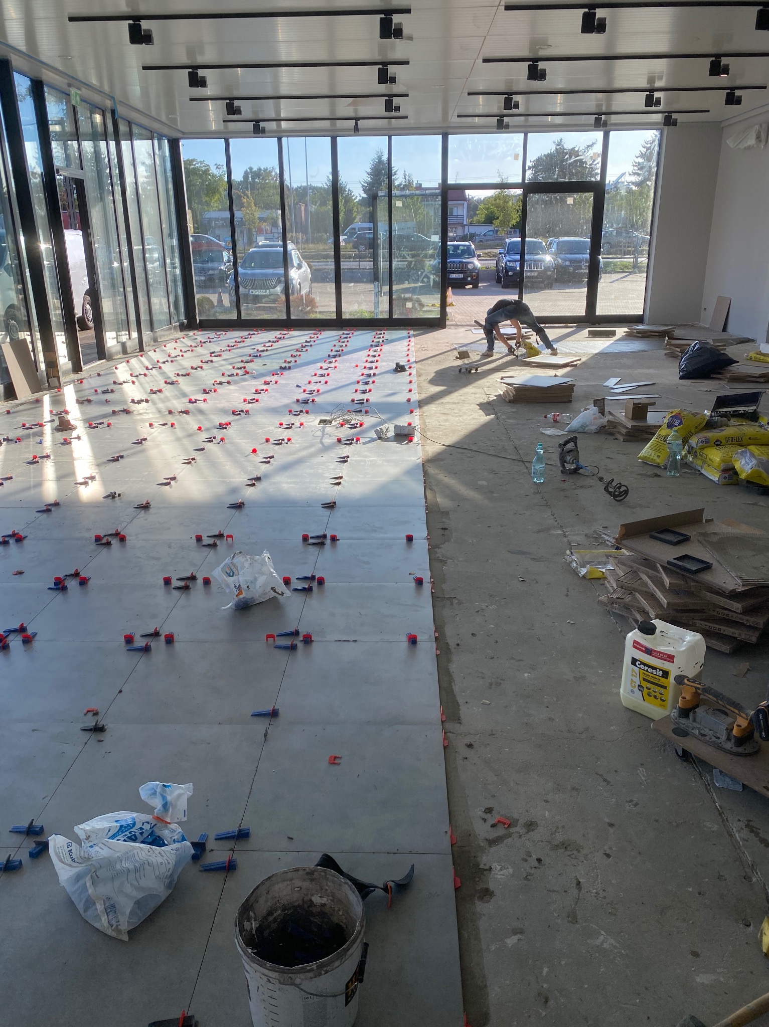 Układanie płytek podłogowych w jasnym pomieszczeniu z dużymi oknami. Widoczne narzędzia, klej i pracownik w trakcie pracy. Częściowo ułożone płytki z systemem poziomowania.