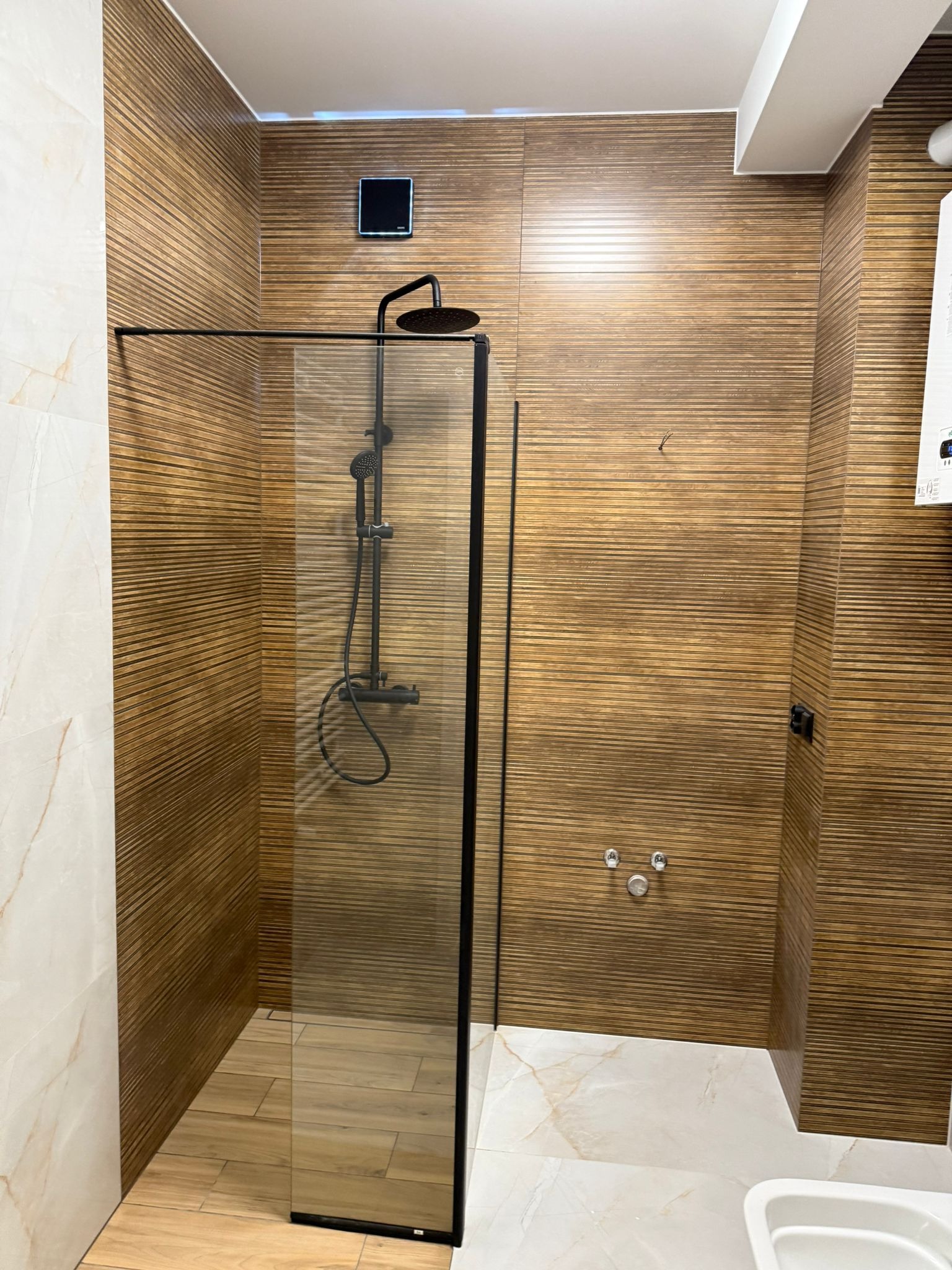Nowoczesna kabina prysznicowa z czarną armaturą i ścianami wyłożonymi płytkami imitującymi drewno. Podłoga wyłożona płytkami imitującymi drewno i marmur.