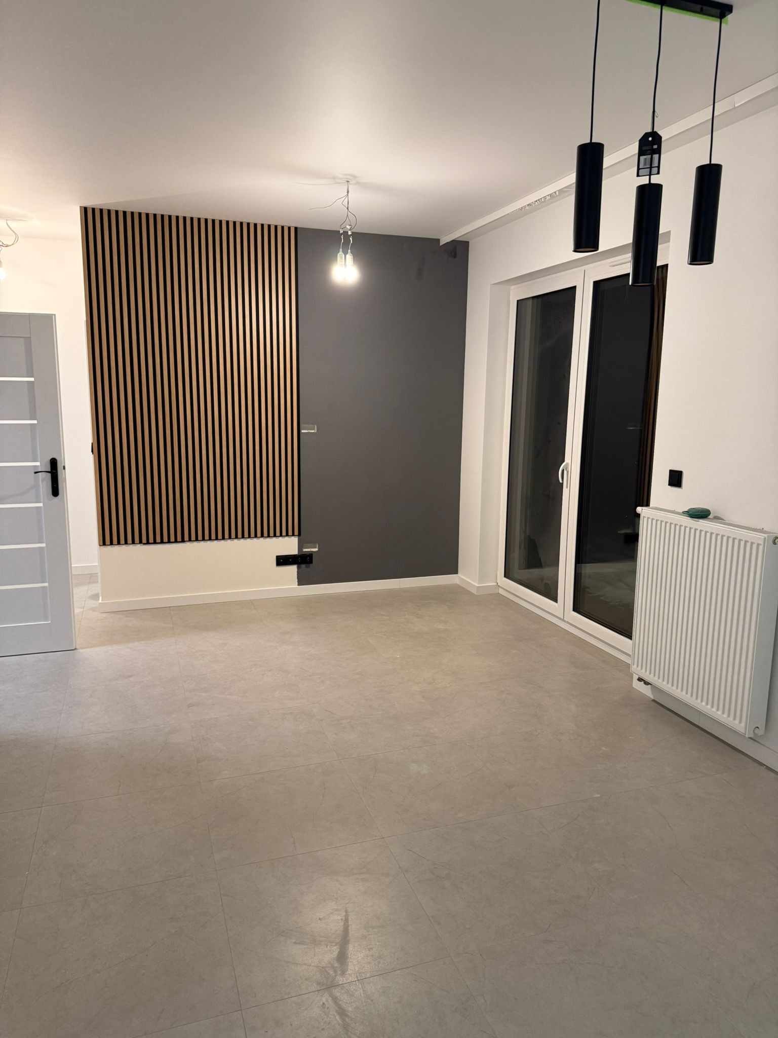 Wykończone wnętrze pokoju z drewnianą ścianą z lameli, szarą ścianą, grzejnikiem i oknem balkonowym. Nowoczesne lampy wiszące.