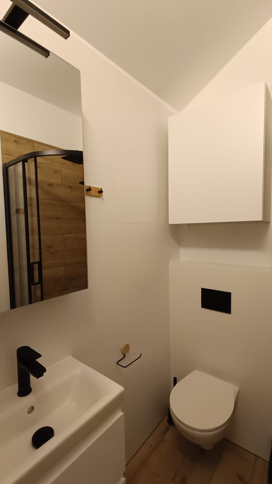 Nowoczesna łazienka w Warszawie z białymi płytkami, czarną armaturą i drewnianą podłogą. W lustrze odbija się kabina prysznicowa z drewnianym wykończeniem.