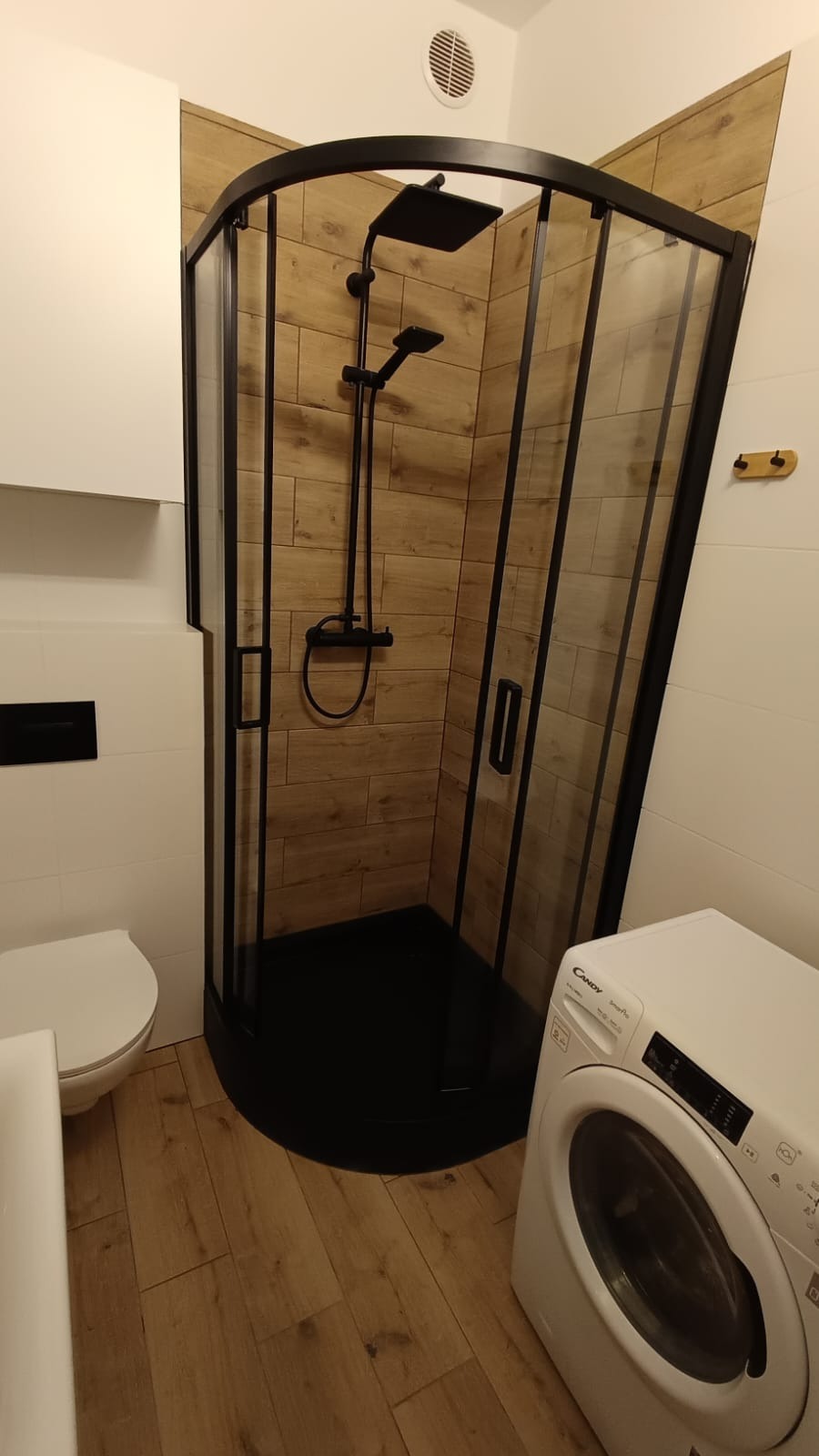 Nowoczesna łazienka po remoncie: kabina prysznicowa z czarną armaturą i drewnopodobnymi płytkami, pralka Candy, biała ceramika. Ujęcie z niskiego kąta.