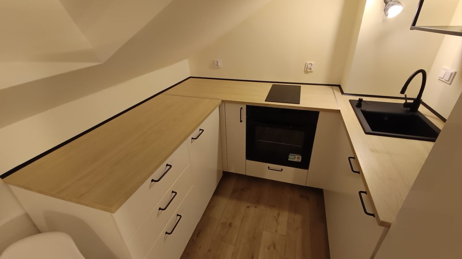 Nowoczesna, jasna kuchnia na poddaszu ze skośnym sufitem. Białe szafki z drewnianym blatem, czarny zlew i bateria. Minimalistyczny design, oświetlenie punktowe.