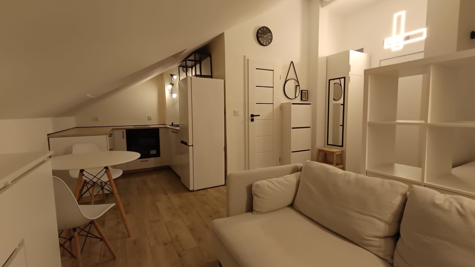 Jasne, minimalistyczne wnętrze poddasza z drewnianą podłogą, białymi meblami i neonowym krzyżem na ścianie. Widoczny aneks kuchenny i część wypoczynkowa.