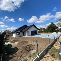 Marek Pawłowski - Nowoczesny dom jednorodzinny z szarą kostką brukową na podjeździe, widok z góry na tle błękitnego nieba z chmurami. W tle traktor i teren w trakcie prac ziemnych.