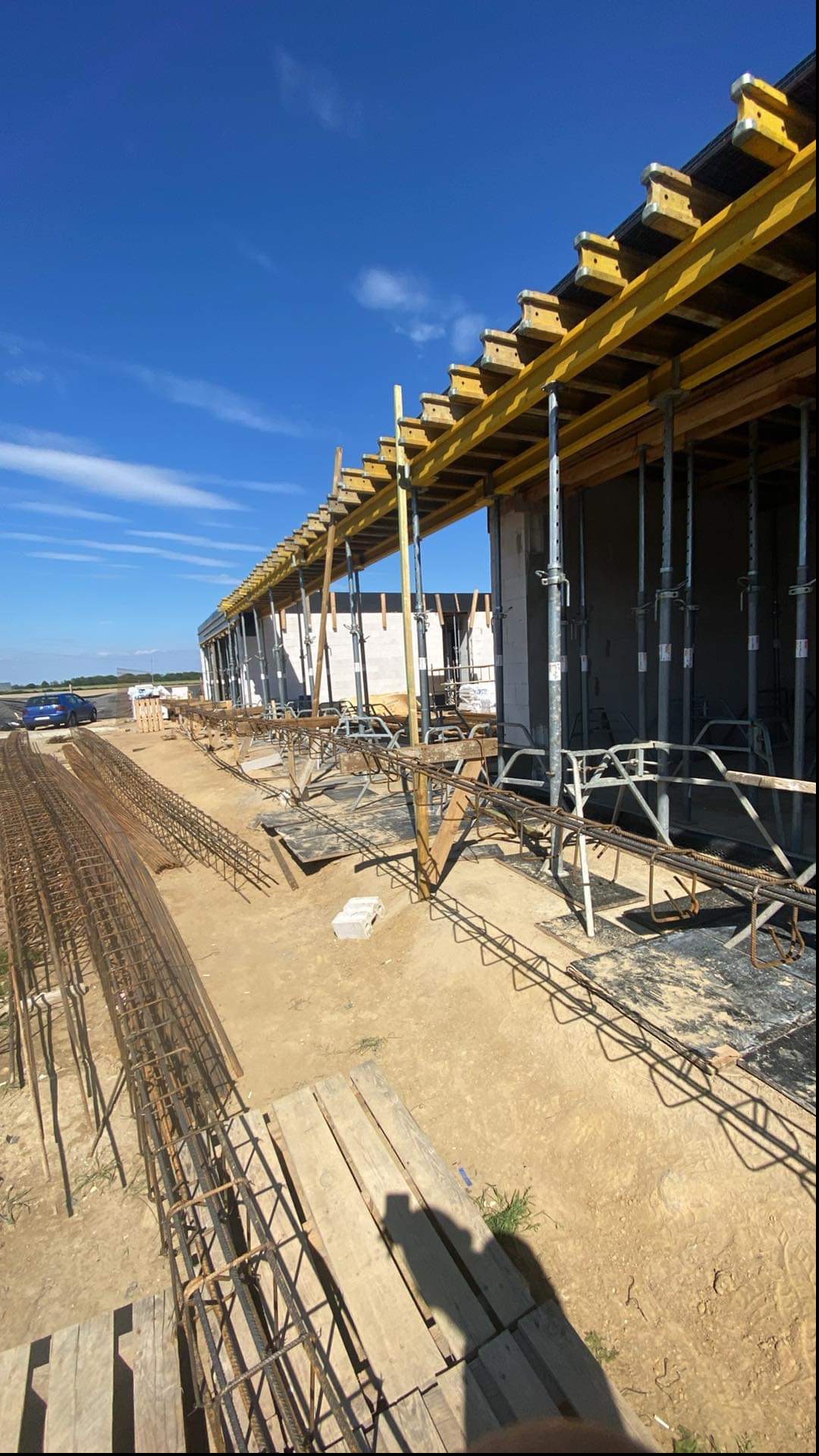 Wykop pod fundamenty z widocznym zbrojeniem i szalunkiem, przygotowany do zalania betonem. W tle ściany budynku w trakcie wznoszenia i błękitne niebo.