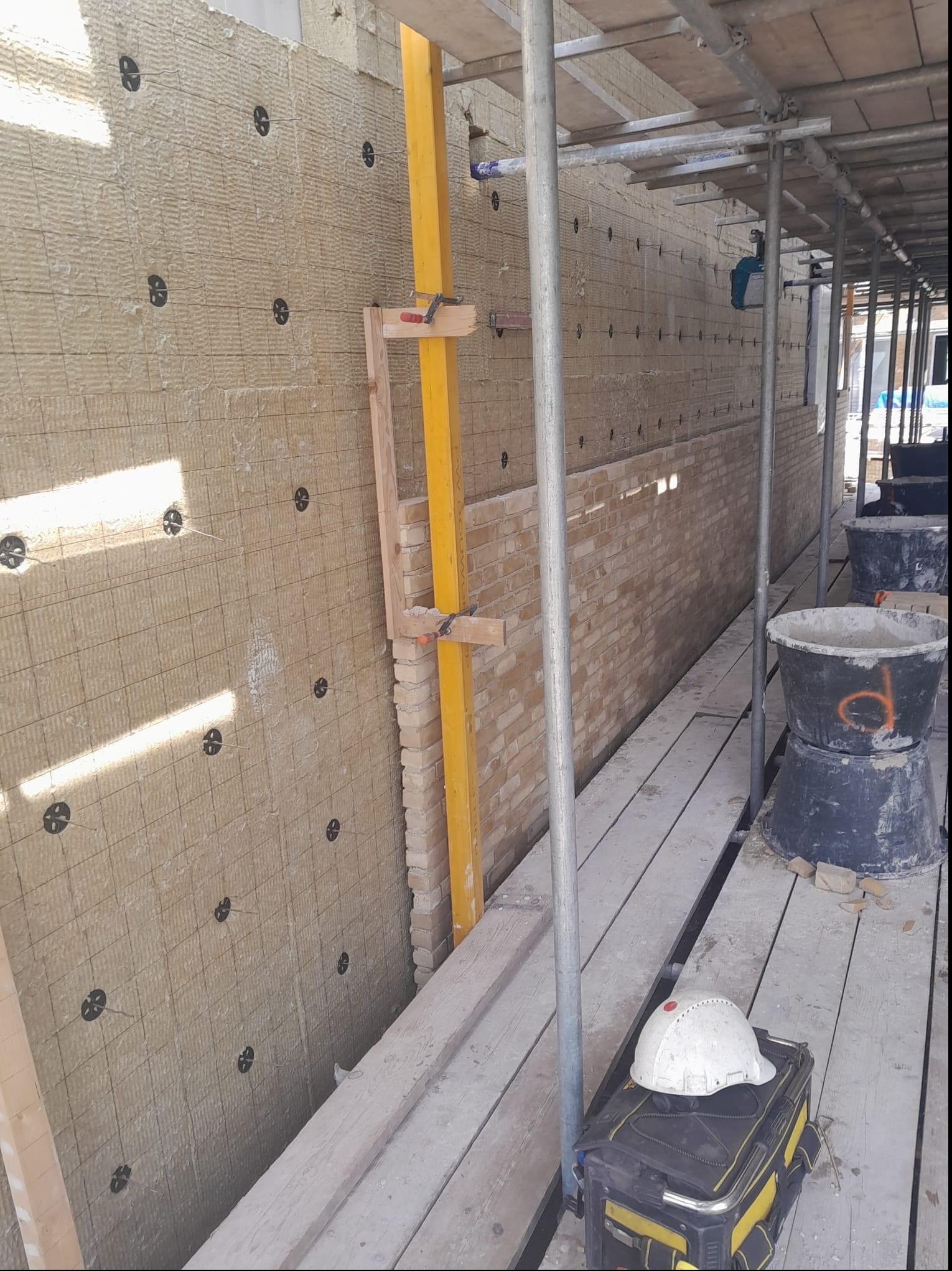 Ocieplanie ściany zewnętrznej wełną mineralną, widoczne rusztowanie, narzędzia i fragment muru z cegły. Prace na wysokości.