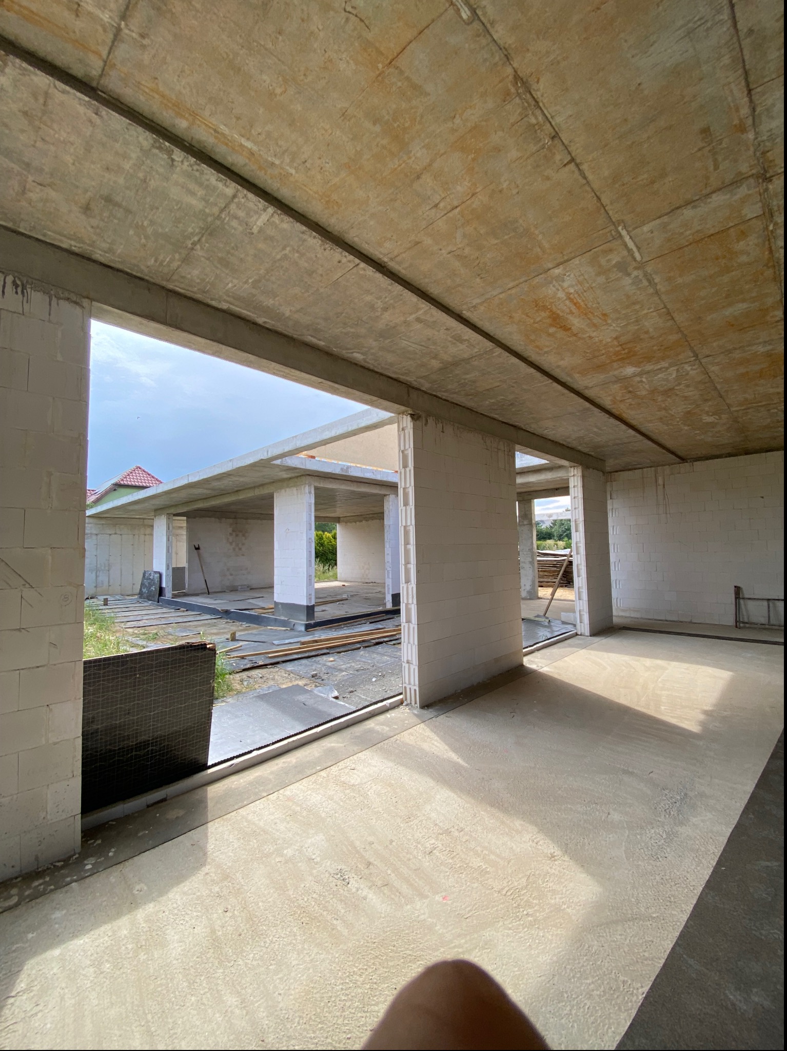 Surowy stan budynku w trakcie budowy: widoczne ściany z bloczków, betonowy strop i podłoga. W tle widok na kolejną część budynku i zieleń.
