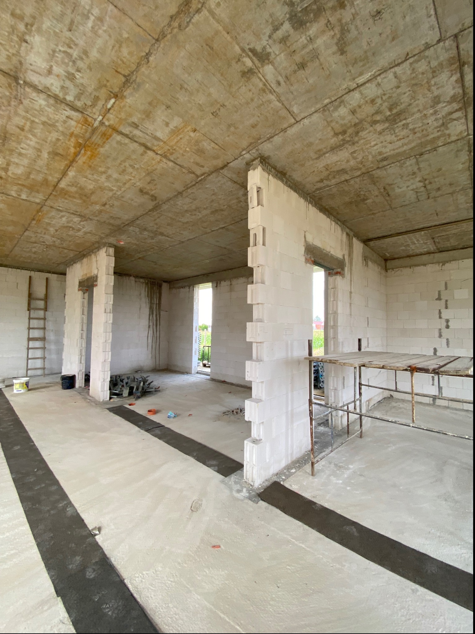 Surowy stan wnętrza budynku w trakcie budowy: ściany z białych bloczków, betonowy strop, widoczne narzędzia i drabina. W tle zieleń za oknem.