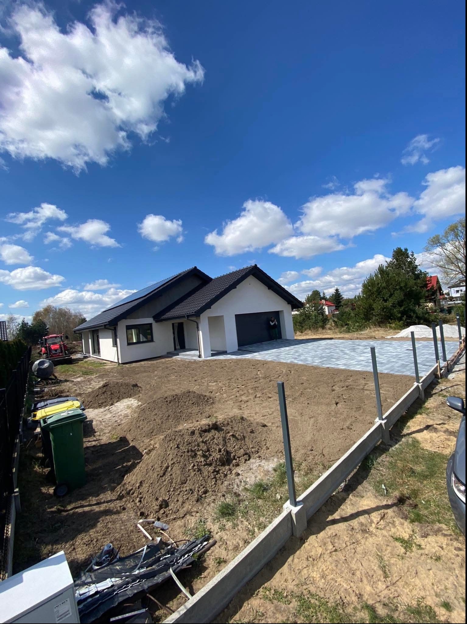 Nowoczesny dom jednorodzinny z szarą kostką brukową na podjeździe, widok z góry na tle błękitnego nieba z chmurami. W tle traktor i teren w trakcie prac ziemnych.