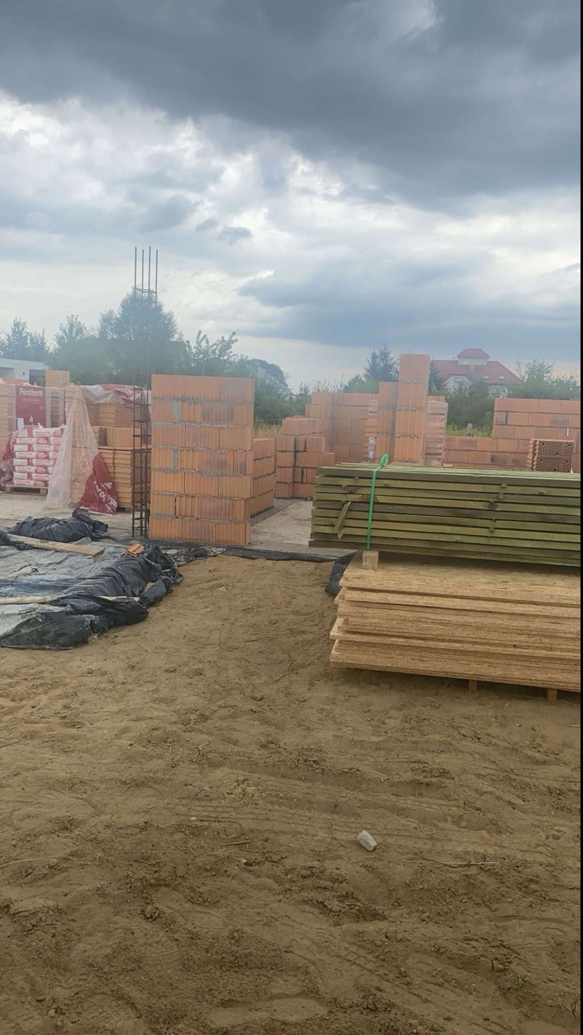Budowa domu w trakcie: cegły Porotherm ułożone w mur, drewniane deski i zbrojenie na placu budowy pod pochmurnym niebem.
