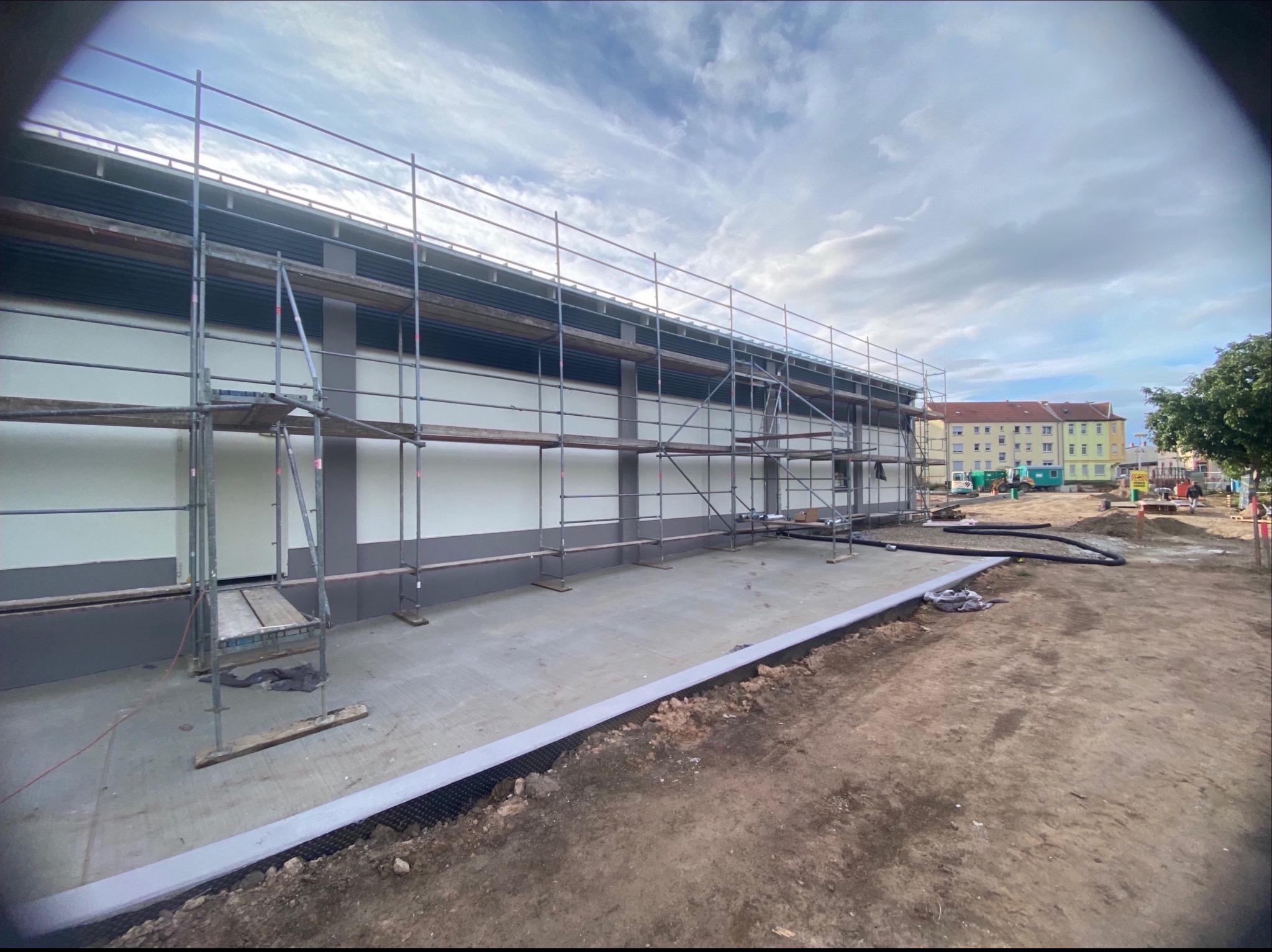 Budynek w trakcie prac elewacyjnych z rusztowaniem, widoczne przygotowane fundamenty i częściowo ocieplona ściana. W tle budynki mieszkalne i pochmurne niebo.