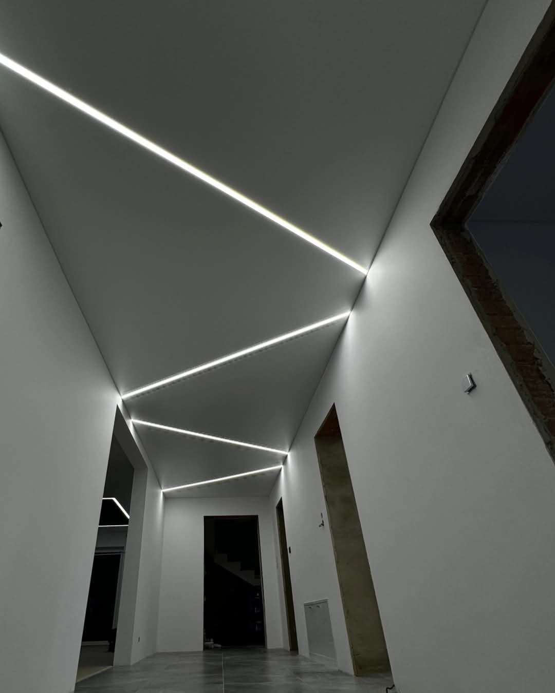 Korytarz z białymi ścianami i geometrycznie ułożonymi, wpuszczonymi liniami świetlnymi LED w suficie, widoczne surowe wykończenie ościeżnicy drzwiowej z cegłą.