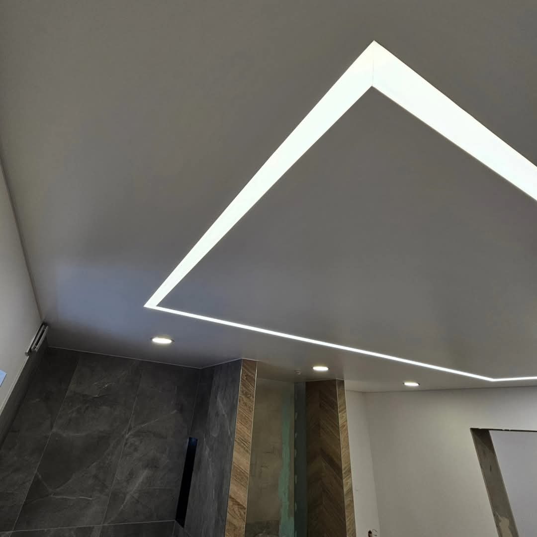 Nowoczesny sufit z oświetleniem LED w kształcie kwadratu, widoczne fragmenty ściany z ciemnymi płytkami imitującymi marmur i drewnianą okładziną ułożoną w jodełkę.