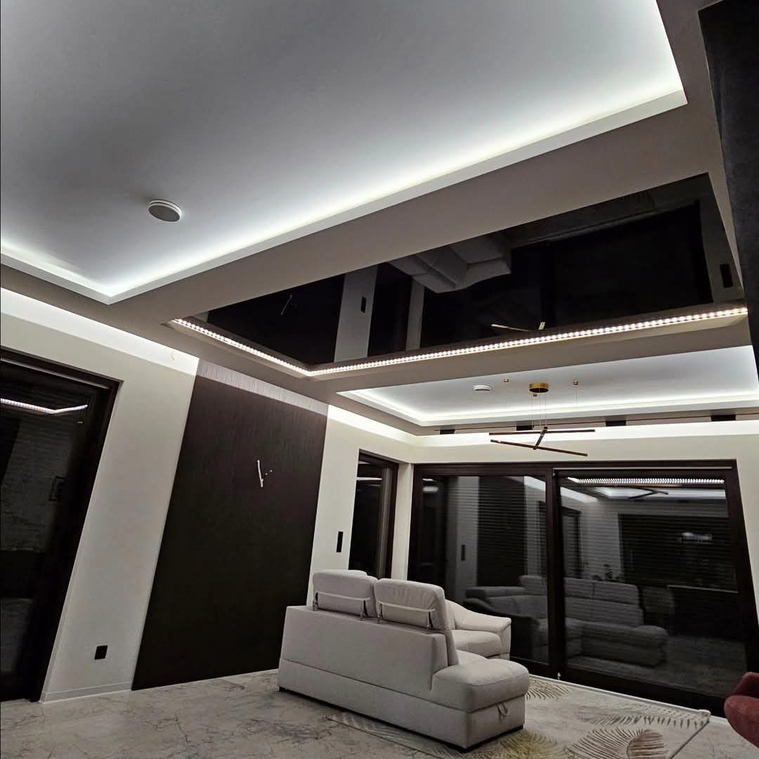 Nowoczesny salon z czarnym, lustrzanym sufitem podwieszanym, oświetlonym taśmami LED, odbijającym sofę i elementy wystroju.