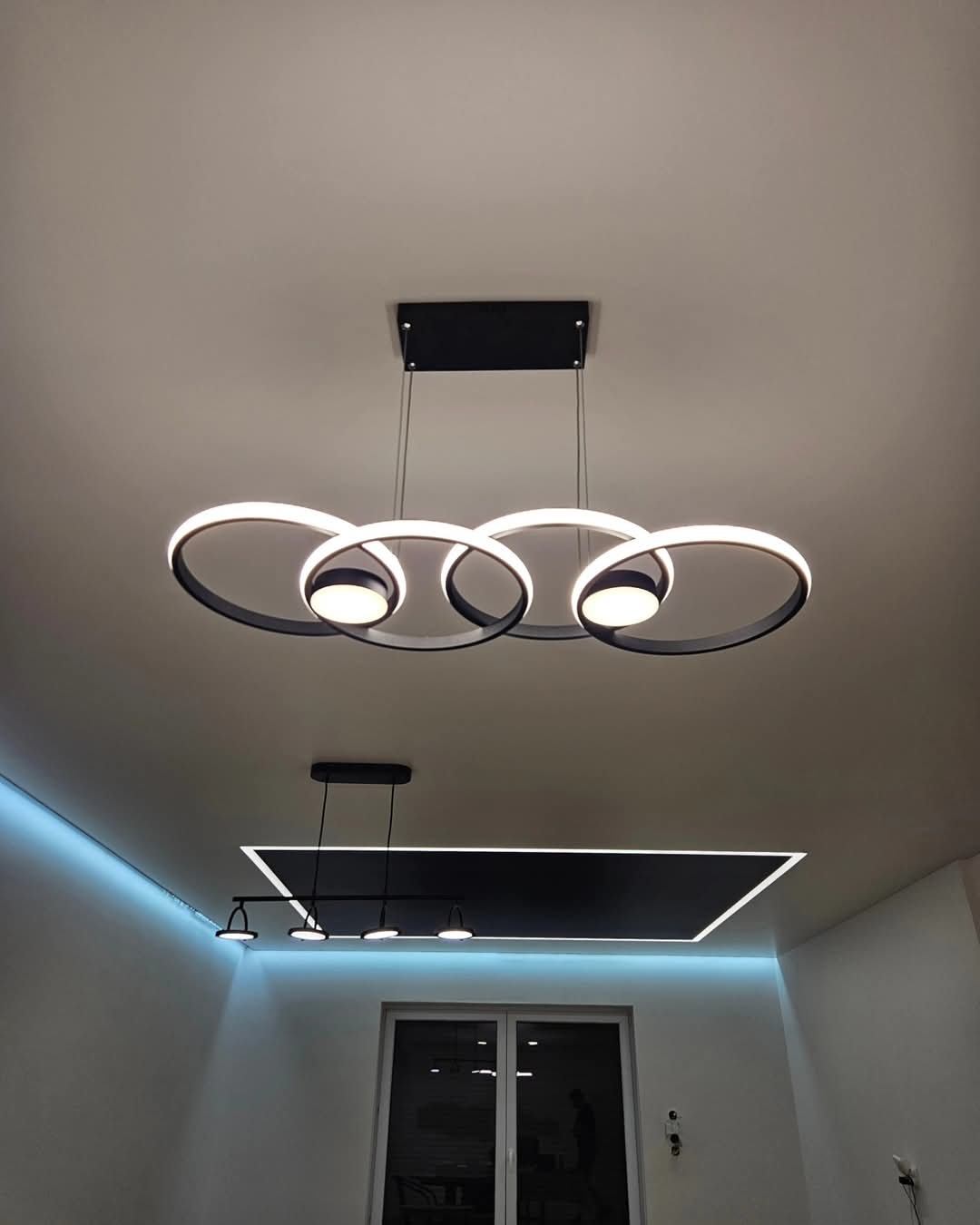 Nowoczesny sufit z designerskim oświetleniem: okrągły żyrandol z czarnymi obręczami i białym światłem LED, dodatkowe oświetlenie w postaci wpuszczanej czarnej ramy i niebieskiej listwy LED wzdłuż...