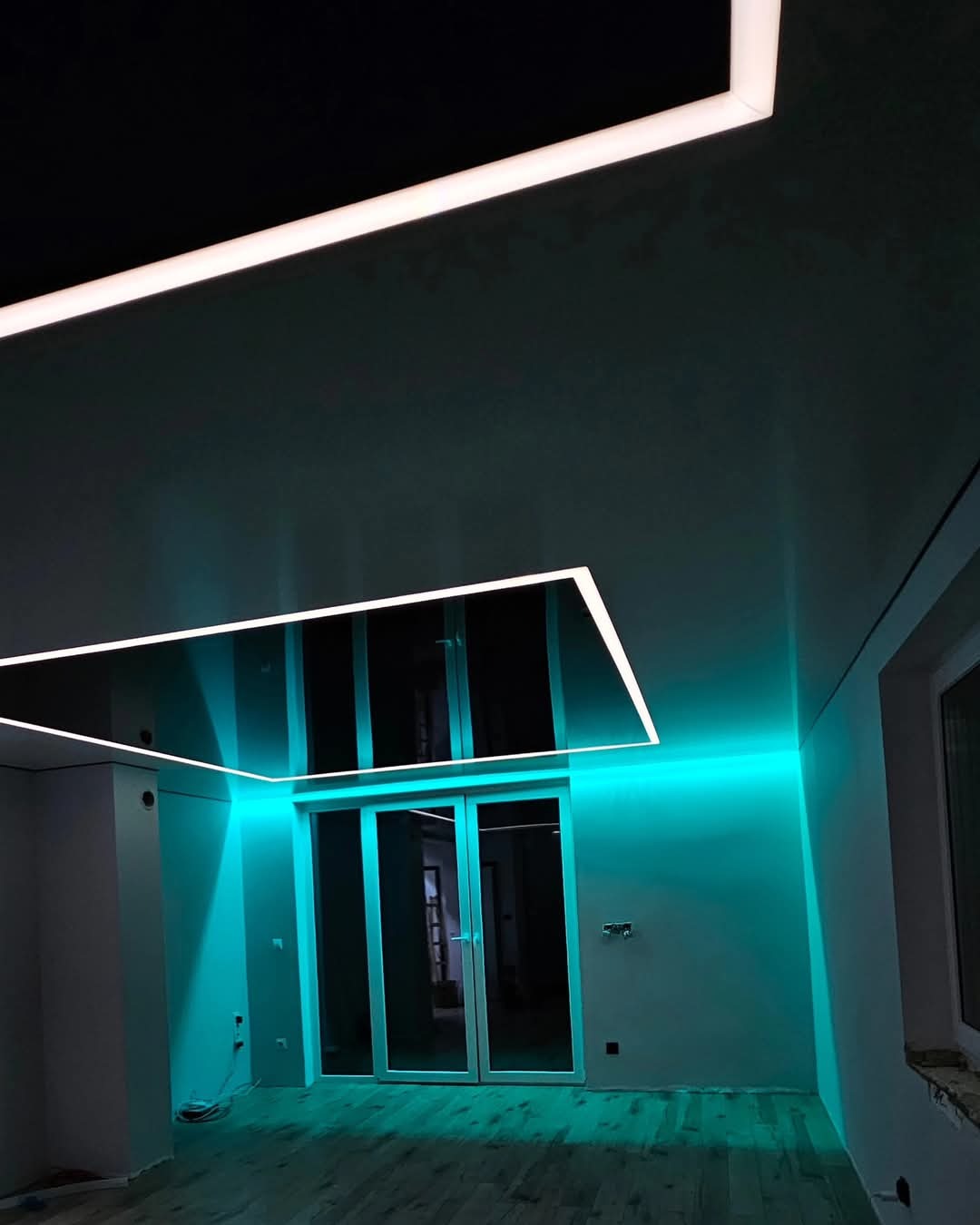 Nowoczesny pokój z czarnym, błyszczącym sufitem podwieszanym z oświetleniem LED w kształcie prostokąta i białym paskiem LED na górze, turkusowe podświetlenie ściany nad szklanymi drzwiami.