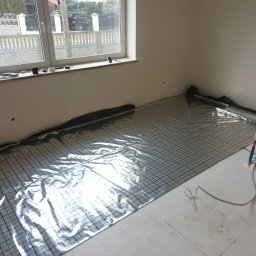 Wod-Gaz Kamil Rosiak - Przygotowanie do montażu ogrzewania podłogowego: mata grzewcza z folią aluminiową i siatką w pomieszczeniu z oknem, widoczne narzędzia i rury.