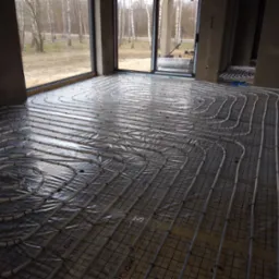 Rozłożona instalacja ogrzewania podłogowego na siatce, rury w kształcie spirali, widoczne okna i surowy stan wykończenia wnętrza.