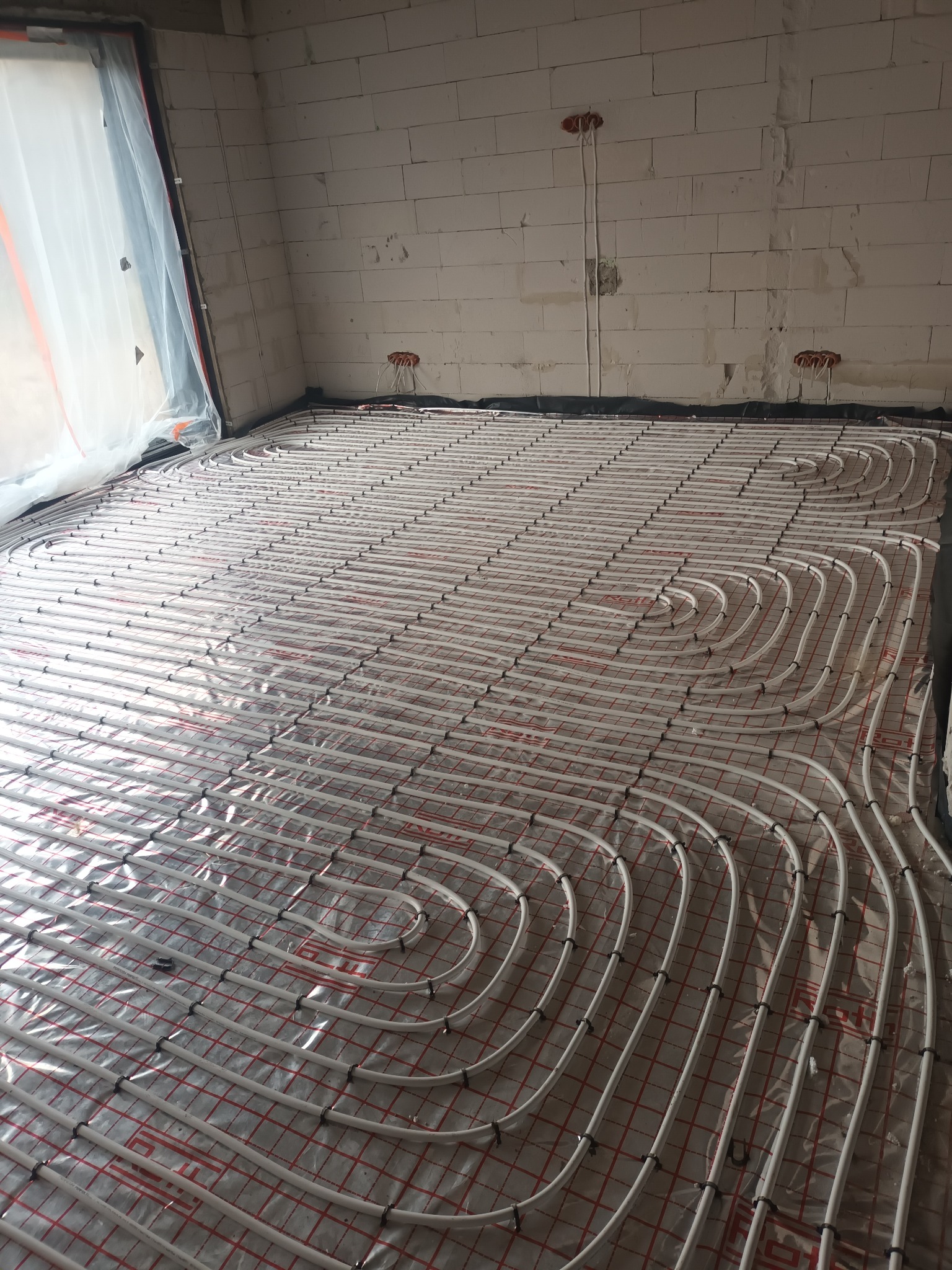 Białe rury ogrzewania podłogowego ułożone na czerwonej siatce w surowym wnętrzu z białych pustaków, widoczne pętle rur i czarne mocowania.