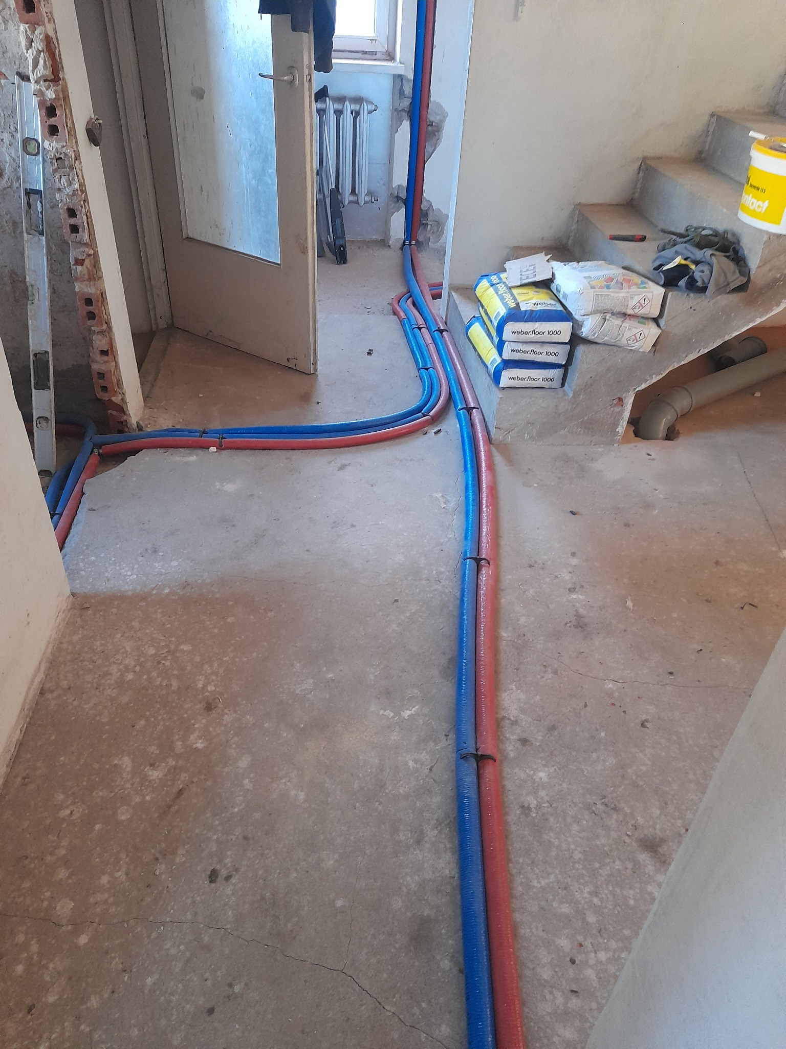 Wnętrze w trakcie remontu z widocznymi, ułożonymi na podłodze, niebieskimi i czerwonymi rurami instalacji wodnej i grzewczej, prowadzącymi od drzwi wejściowych w kierunku schodów, obok których leżą...