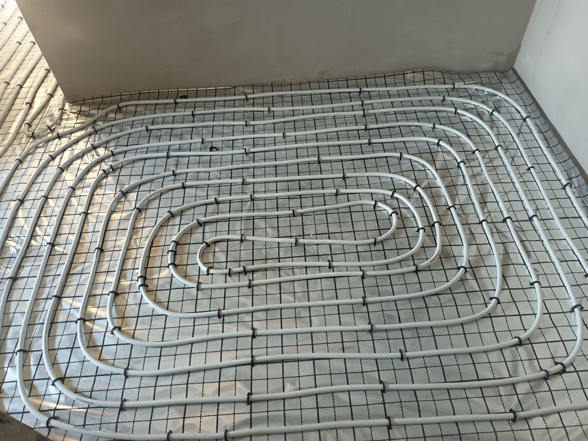 Białe rurki ogrzewania podłogowego ułożone spiralnie na folii z nadrukowaną siatką, przymocowane czarnymi klipsami do podłoża, widok z góry.