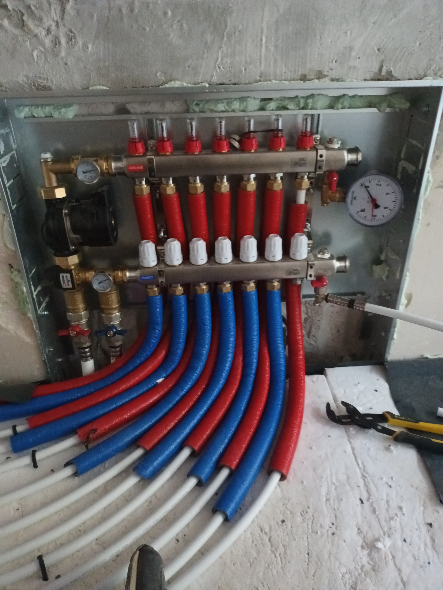 Rozdzielacz ogrzewania podłogowego w metalowej skrzynce, z czerwono-niebieskimi rurami izolowanymi termicznie, widoczne manometry i zawory, surowy stan wykończenia ściany w tle.