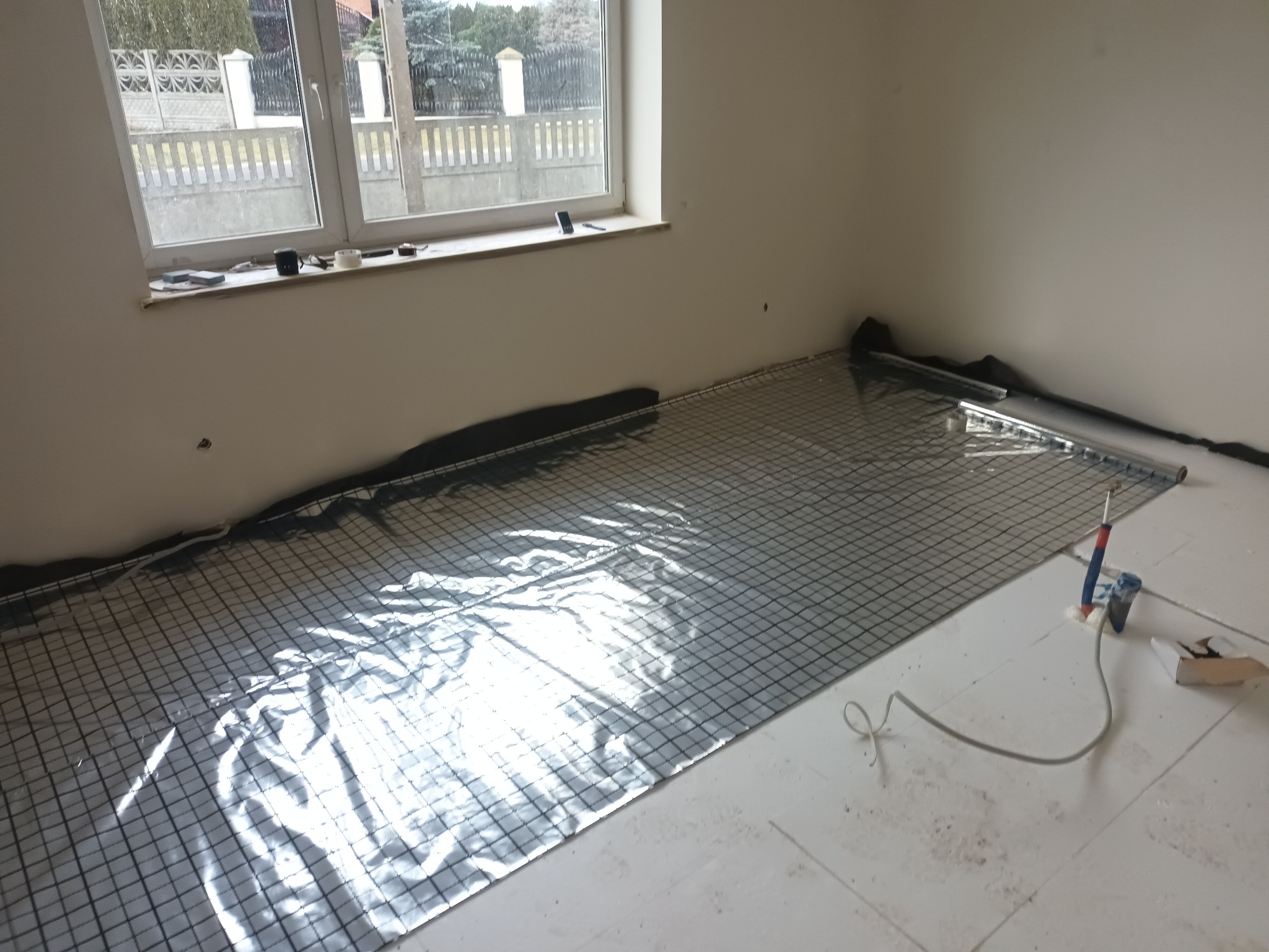 Przygotowanie do montażu ogrzewania podłogowego: mata grzewcza z folią aluminiową i siatką w pomieszczeniu z oknem, widoczne narzędzia i rury.