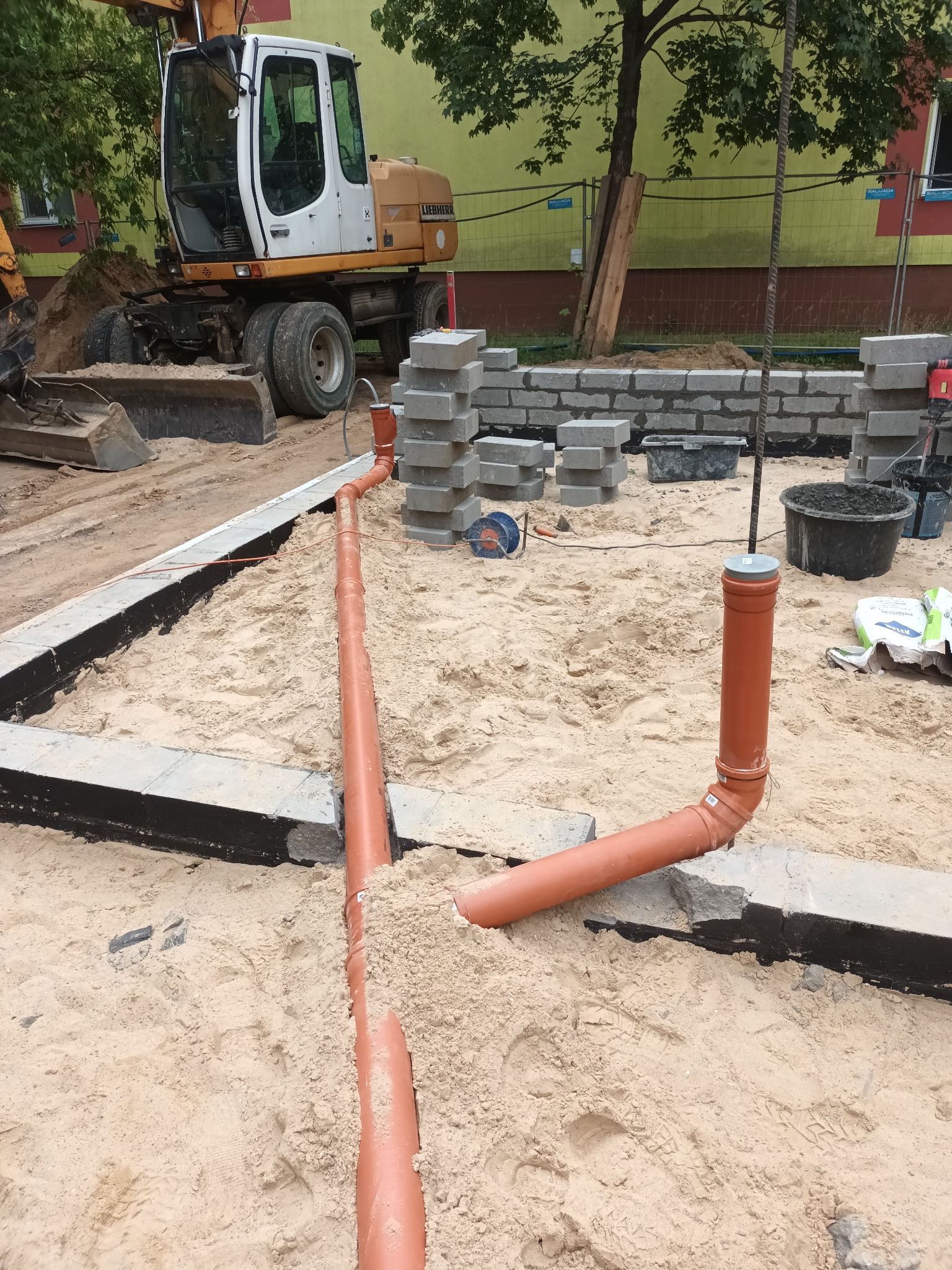 Wykonanie przyłącza kanalizacyjnego z rur PVC w piasku, widoczny wykop z izolacją fundamentów, ułożone bloczki betonowe i koparka Liebherr w tle.
