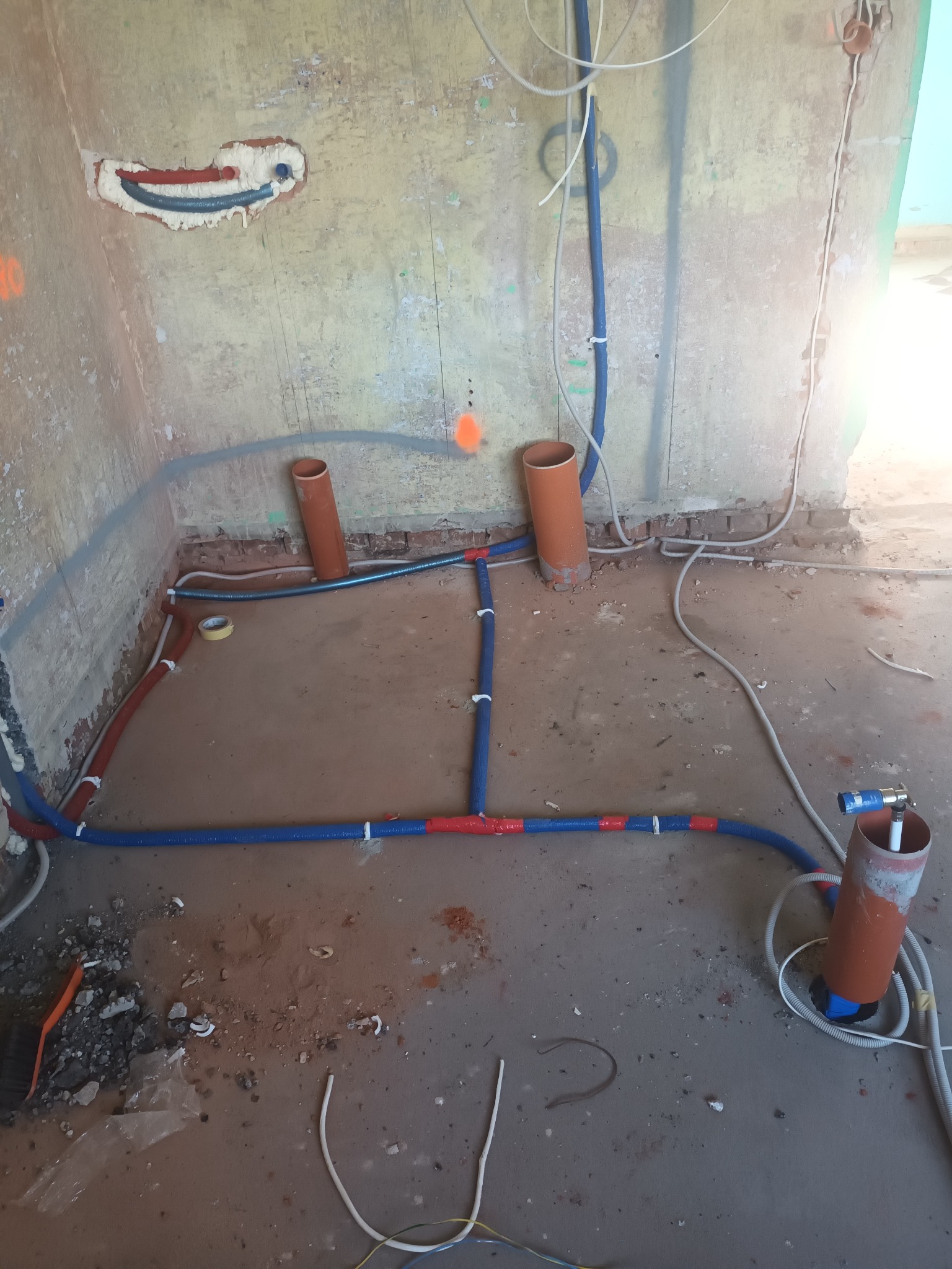 Wnętrze w trakcie remontu z widocznymi, częściowo zamontowanymi rurami instalacji wodnej w kolorze niebiesko-czerwonym, prowadzącymi po podłodze i ścianach, oraz wystającymi pionami kanalizacyjnymi...