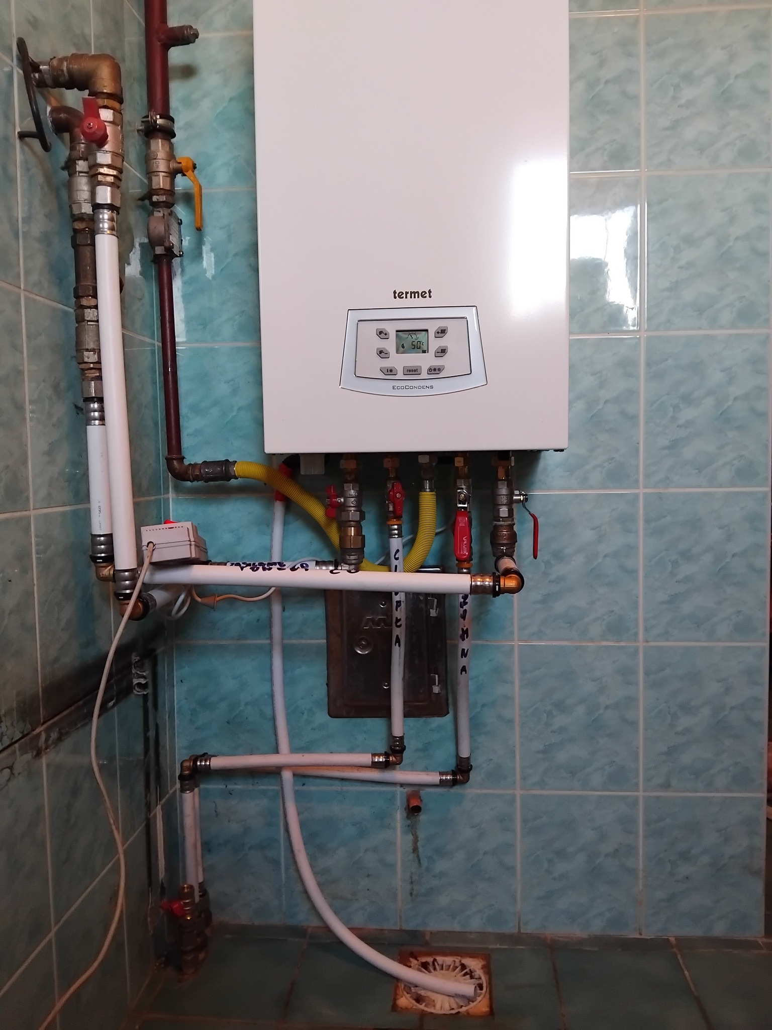 Biały piec gazowy Termet zamontowany na ścianie z niebieskich płytek, widoczne przyłącza gazowe i hydrauliczne, rury z zaworami oraz instalacja odprowadzająca skropliny.