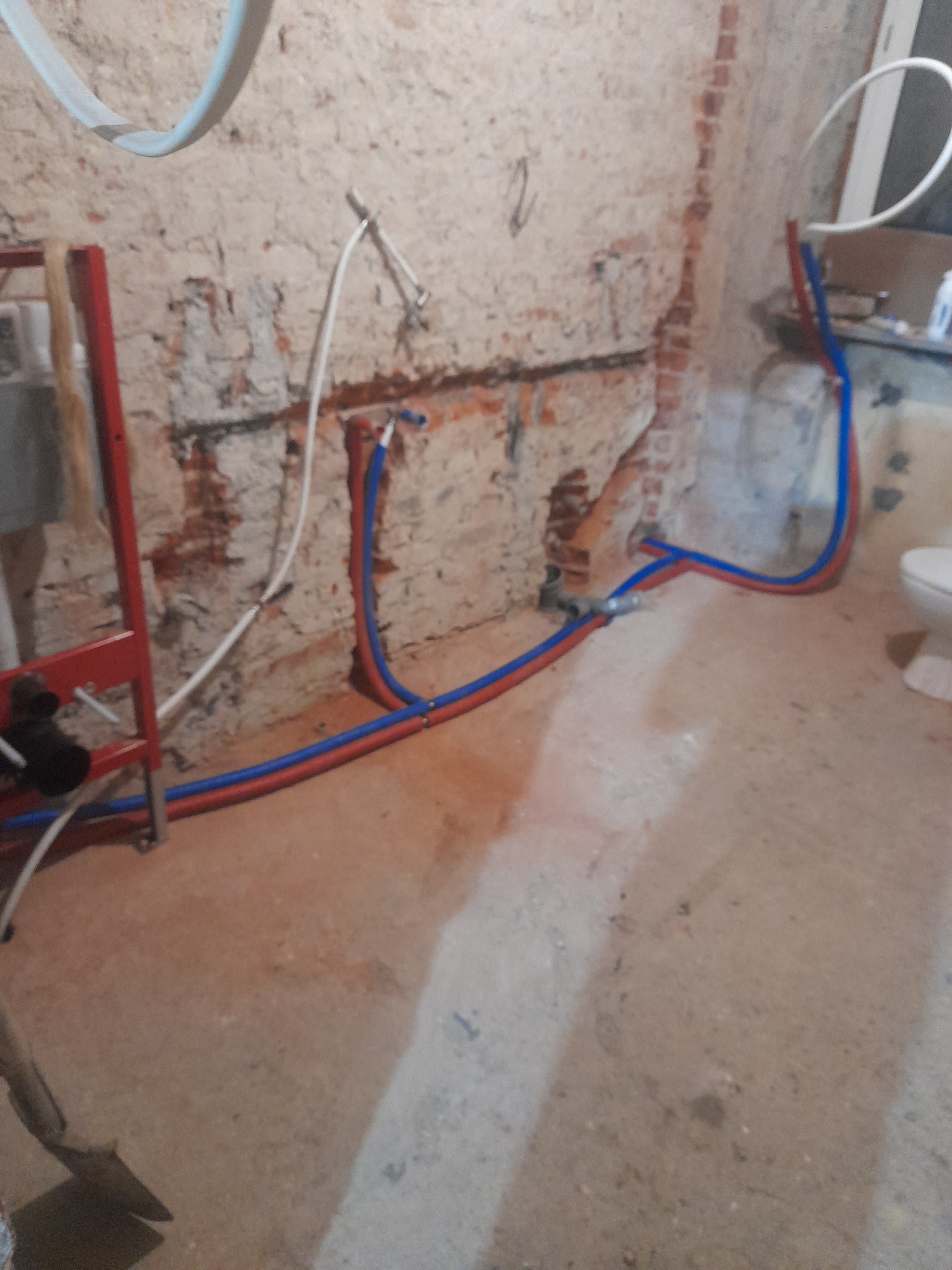Surowe wnętrze łazienki w trakcie remontu, z odsłoniętą cegłą, nowymi rurami PEX w kolorach niebieskim i czerwonym, oraz częściowo zamontowaną stelażą podtynkową WC.