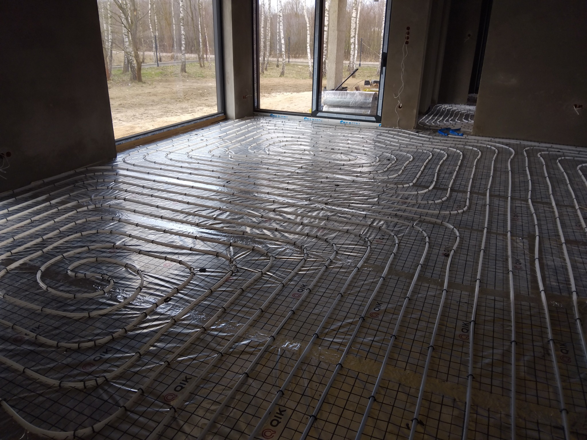 Rozłożona instalacja ogrzewania podłogowego na siatce, rury w kształcie spirali, widoczne okna i surowy stan wykończenia wnętrza.