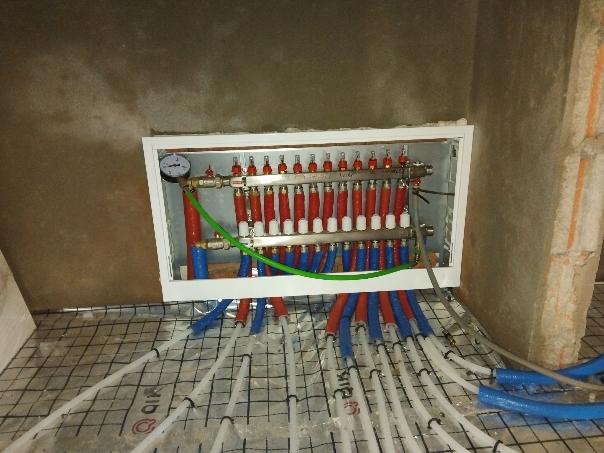 Rozdzielacz ogrzewania podłogowego w skrzynce, z widocznymi rurami czerwonymi i niebieskimi, manometrem, oraz rurami ułożonymi w siatce na podłodze w trakcie montażu.