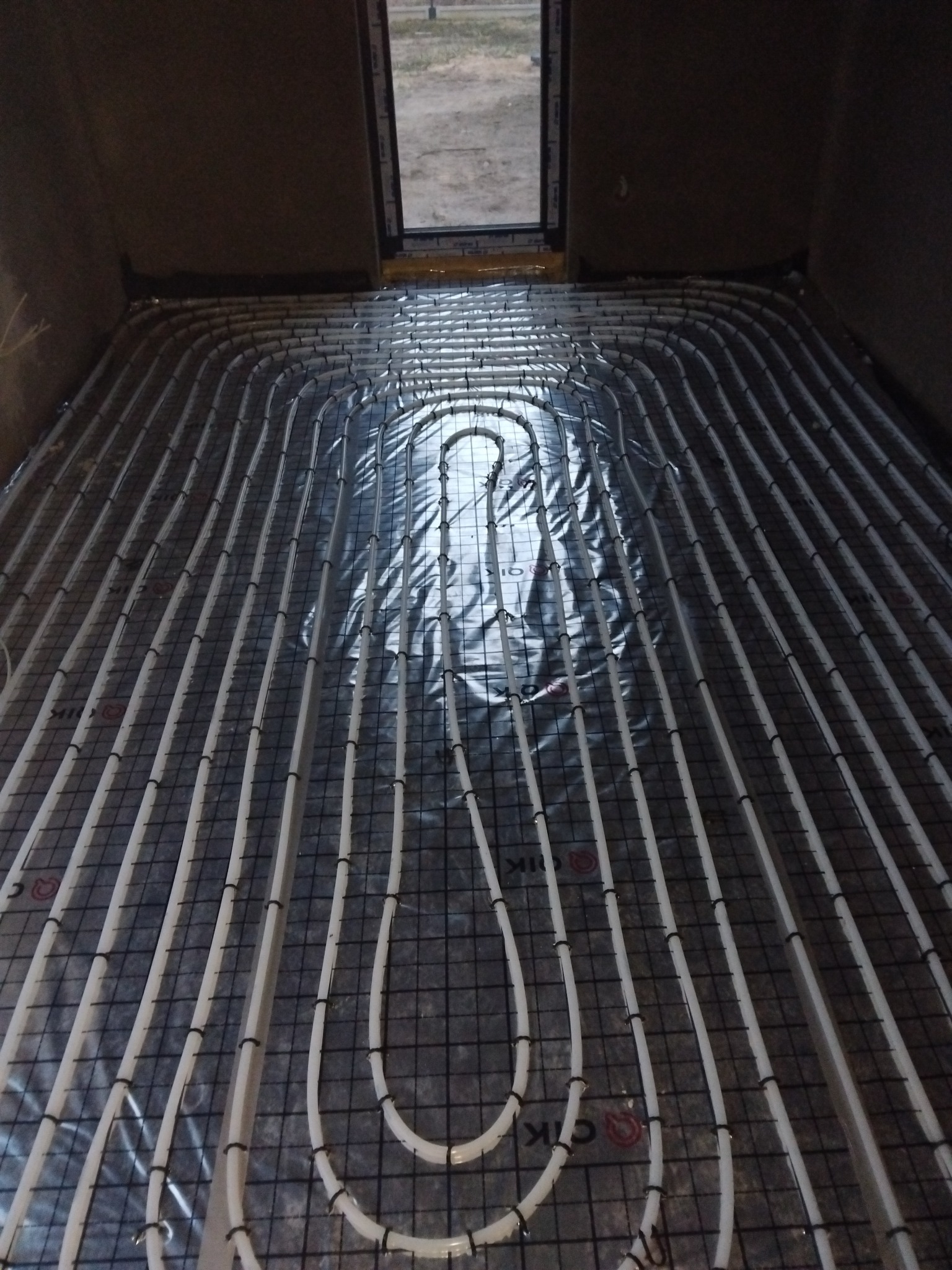 Białe rurki ogrzewania podłogowego ułożone na czarnej siatce, widok z góry w surowym pomieszczeniu z oknem w tle.