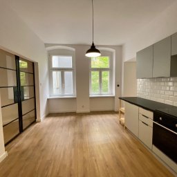 Michał Rostowski - Wykończona kuchnia w jasnych barwach z drewnianą podłogą, szarymi szafkami i czarnym blatem, widok na okno z zielenią za oknem oraz czarne, przeszklone drzwi przesuwne.