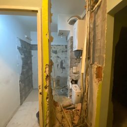 Michał Rostowski - Wnętrze w trakcie demontażu ścianki działowej, odsłonięte przewody elektryczne i instalacja wodna, widoczna łazienka z częściowo położonymi płytkami i bojlerem.